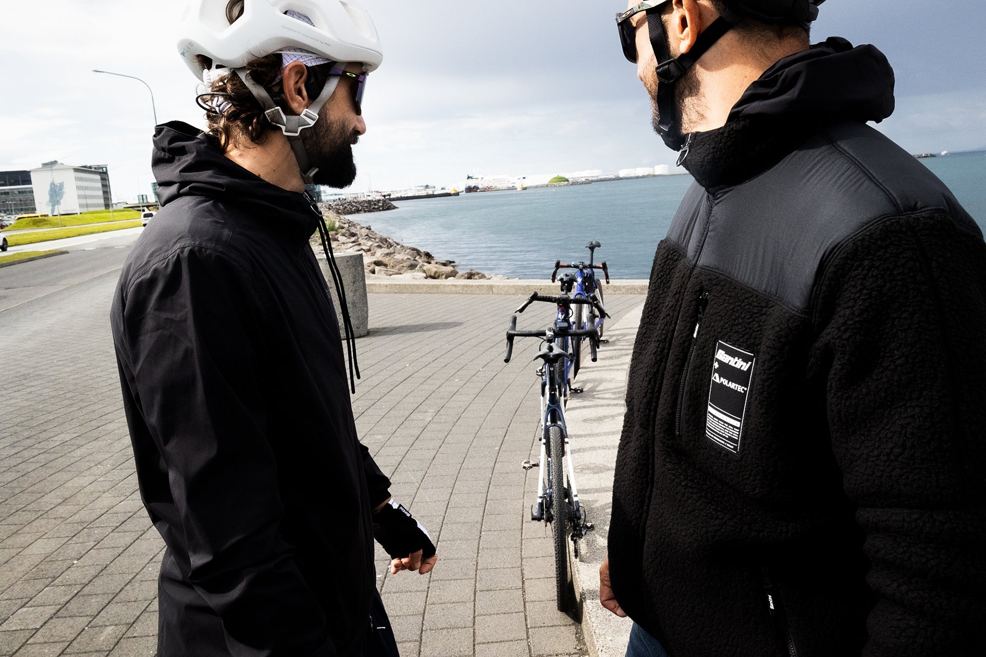 Two men in black jackets and helmets stand facing each other near the water, with a blue bicycle parked on the sidewalk between them, during daytime with overcast sky.
