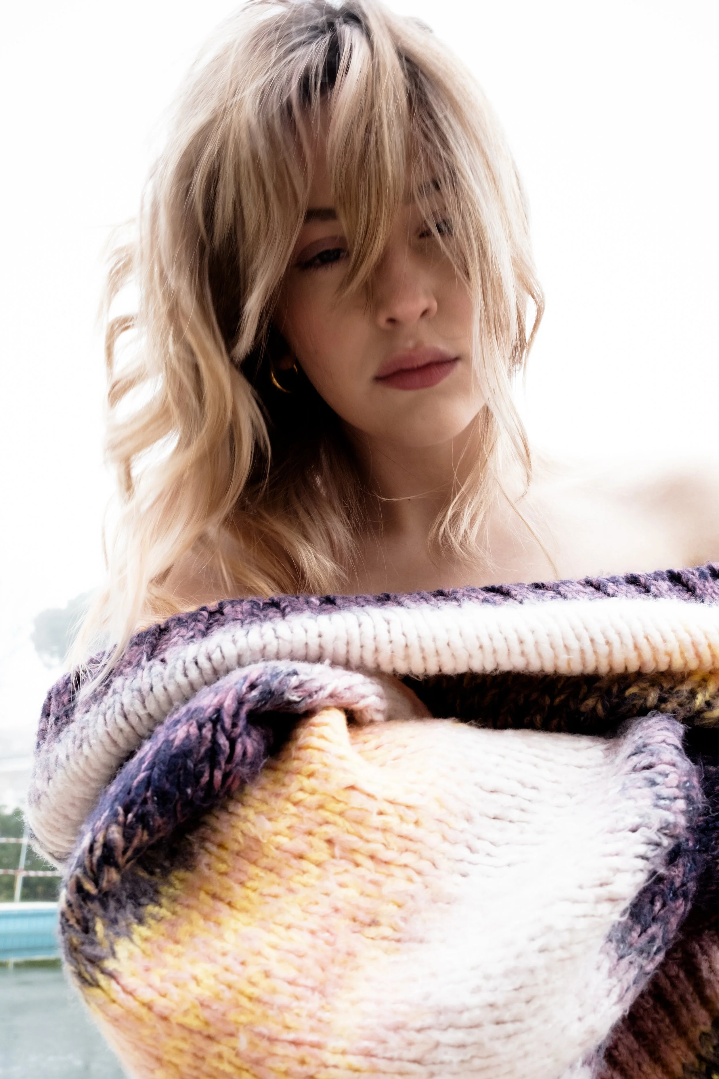 Close-up of a young woman with wavy blonde hair, wearing a multicolored knitted sweater, standing outdoors against a cloudy sky.
