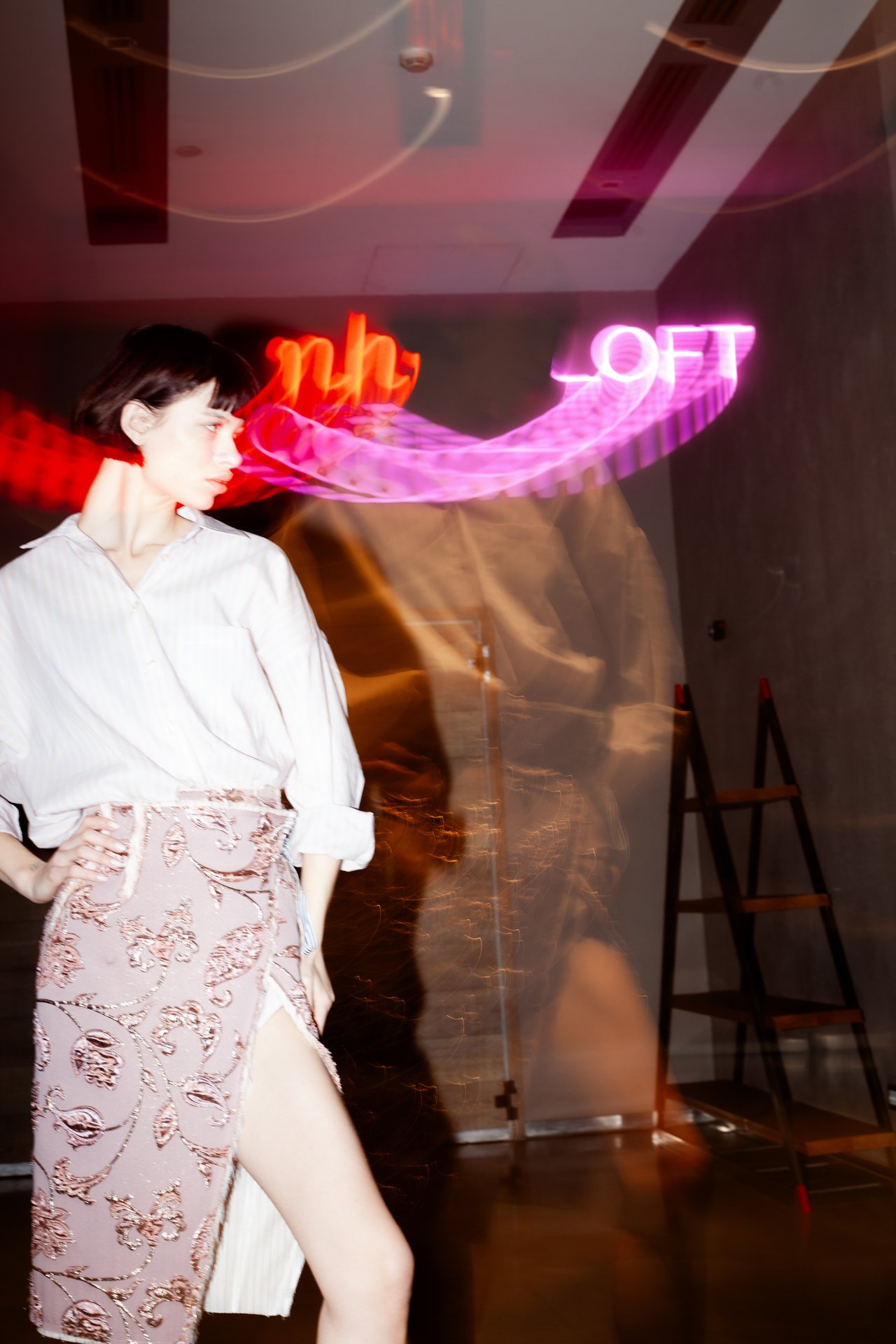 A woman with short dark hair wearing a white shirt and a pink patterned skirt standing in front of a wall with neon signs that read 'Rough' and 'LOFT'. The photo has a blurred, long exposure effect.