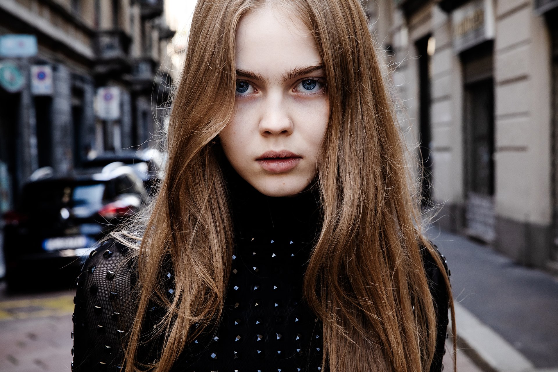 Close-up of a young woman with long red hair, blue eyes, and light skin standing on a city street with buildings and parked cars in the background.