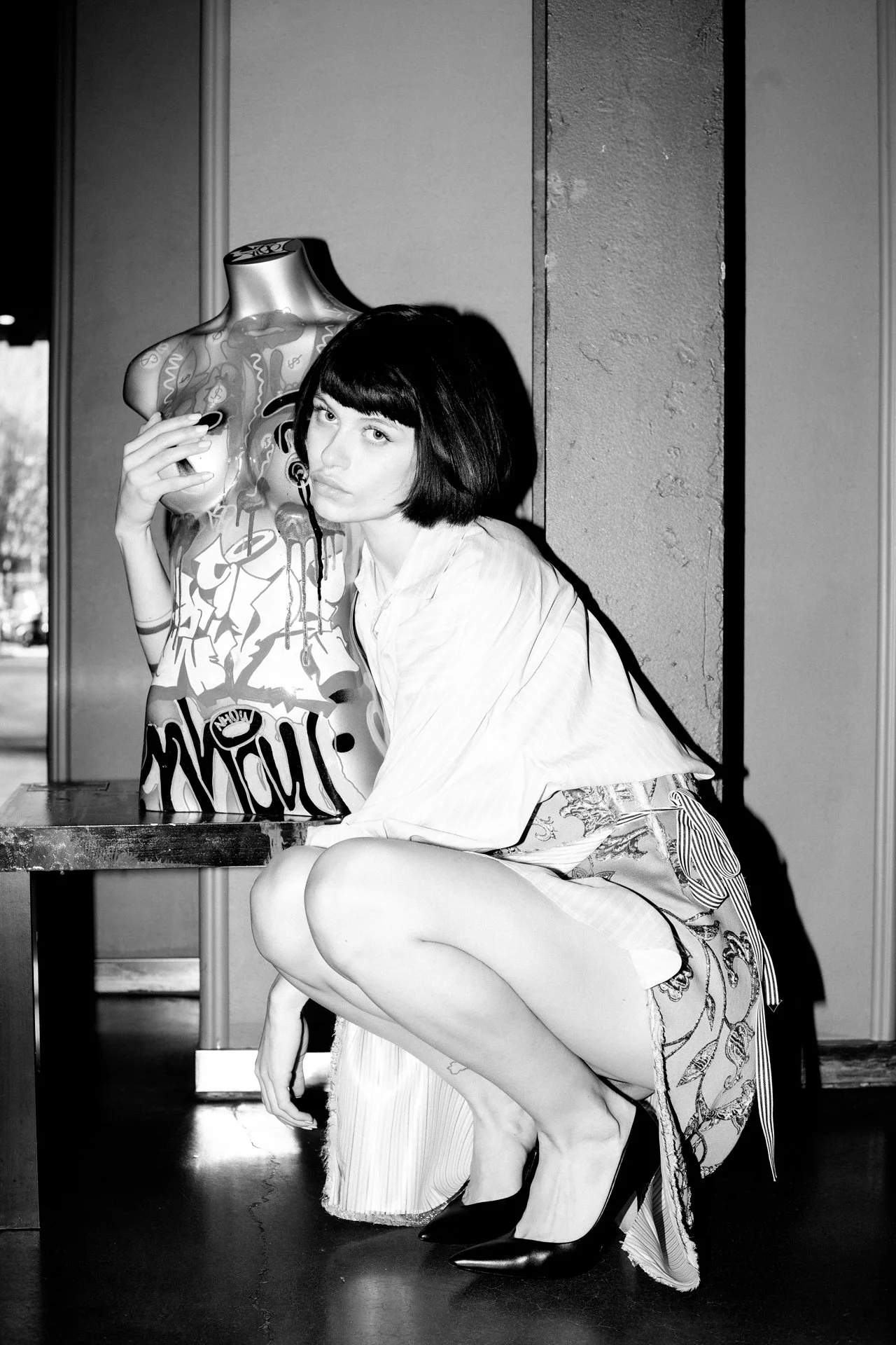 A woman with a short bob haircut crouching next to a life-sized mannequin torso with graffiti-style art, in black and white.