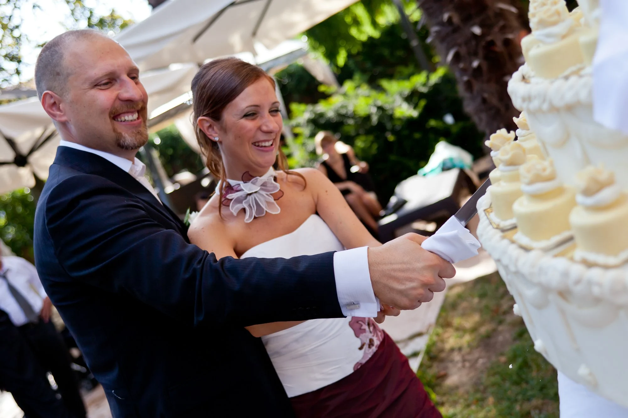 A bride and groom cutting a wedding cake outdoors during their celebration, smiling happily.