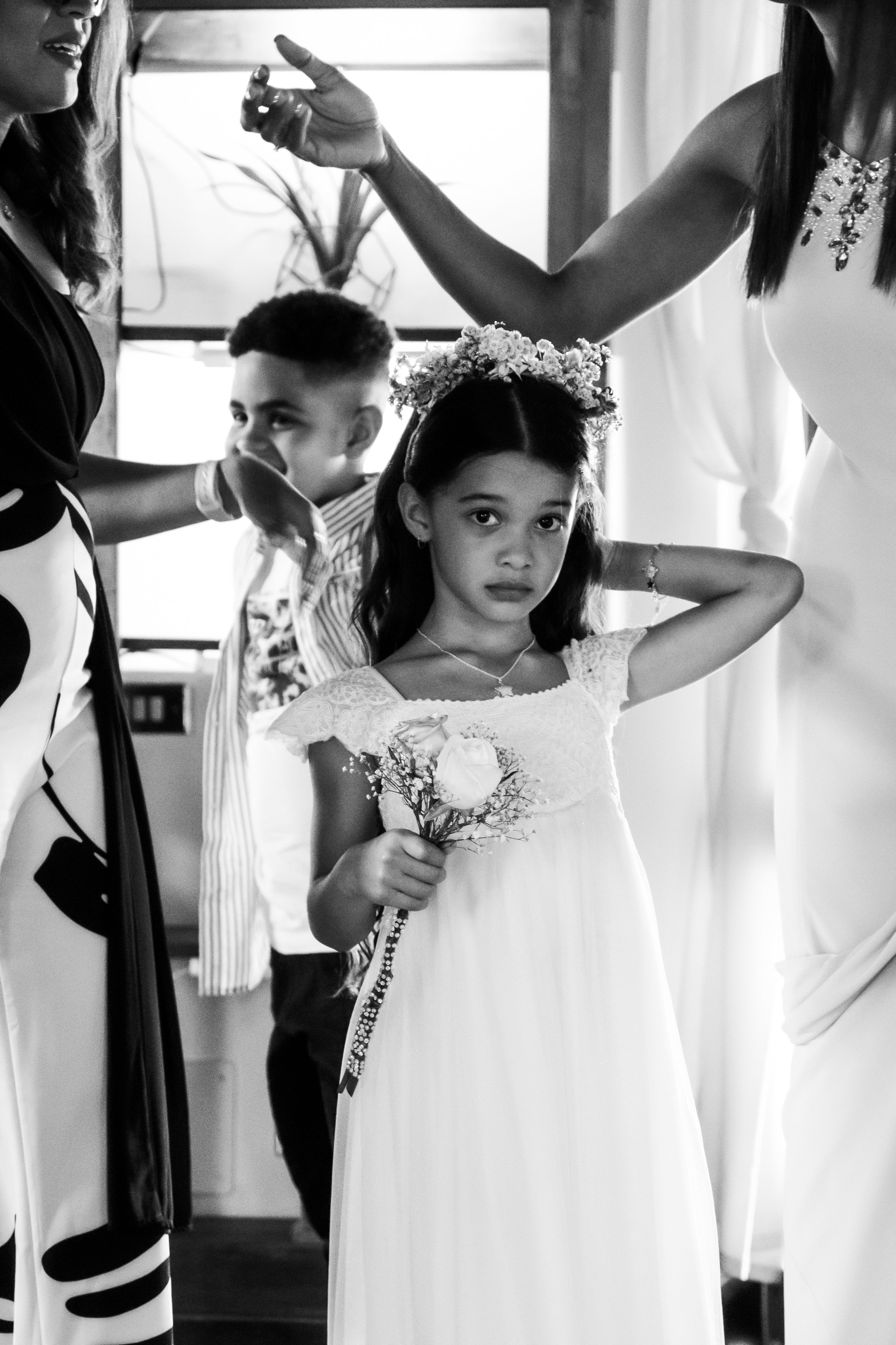 A young girl in a white dress holding a flower, participating in a wedding or similar ceremony, with adults and a boy in the background.