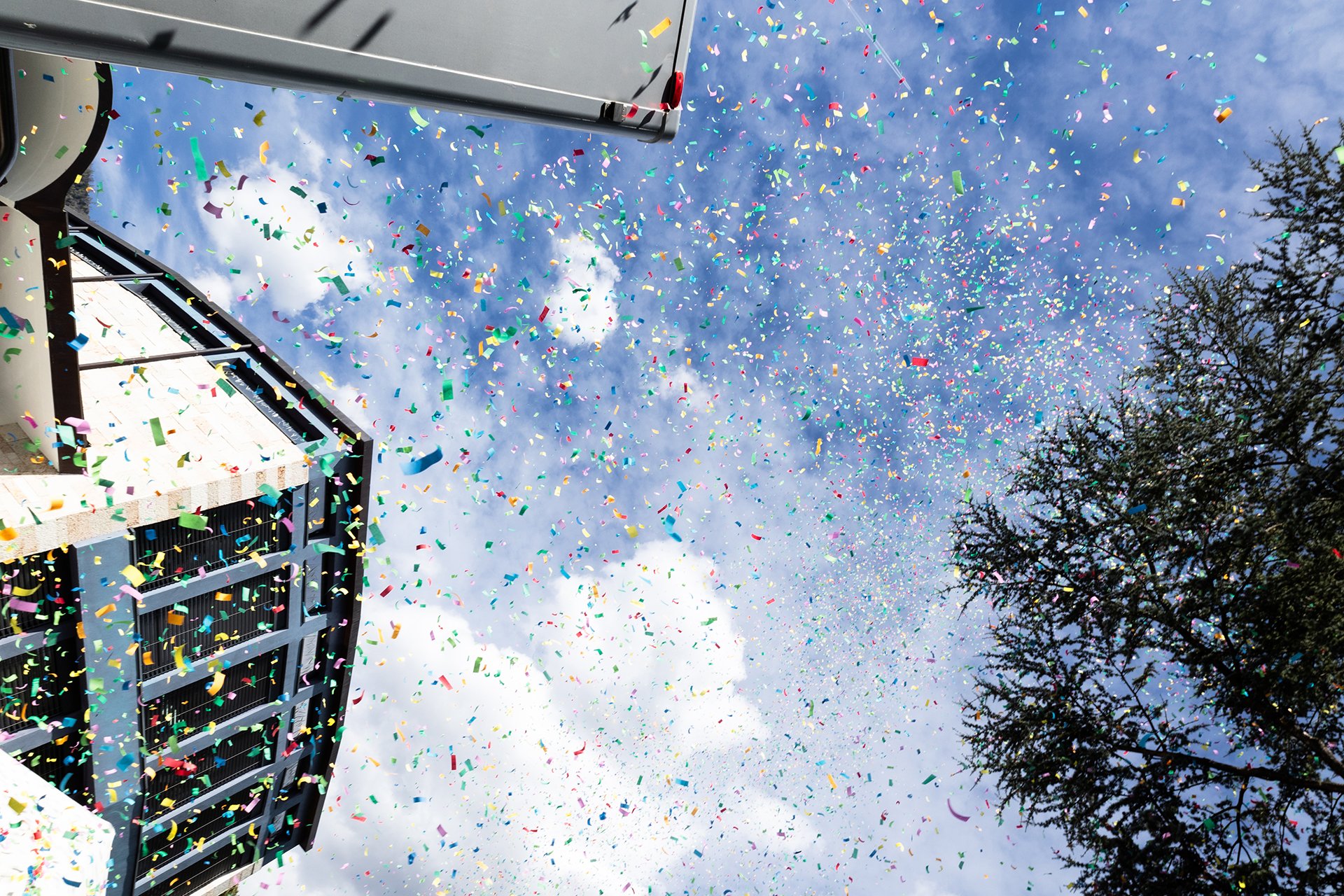 Colorful confetti falling from a high building with a blue sky and some clouds, alongside trees.