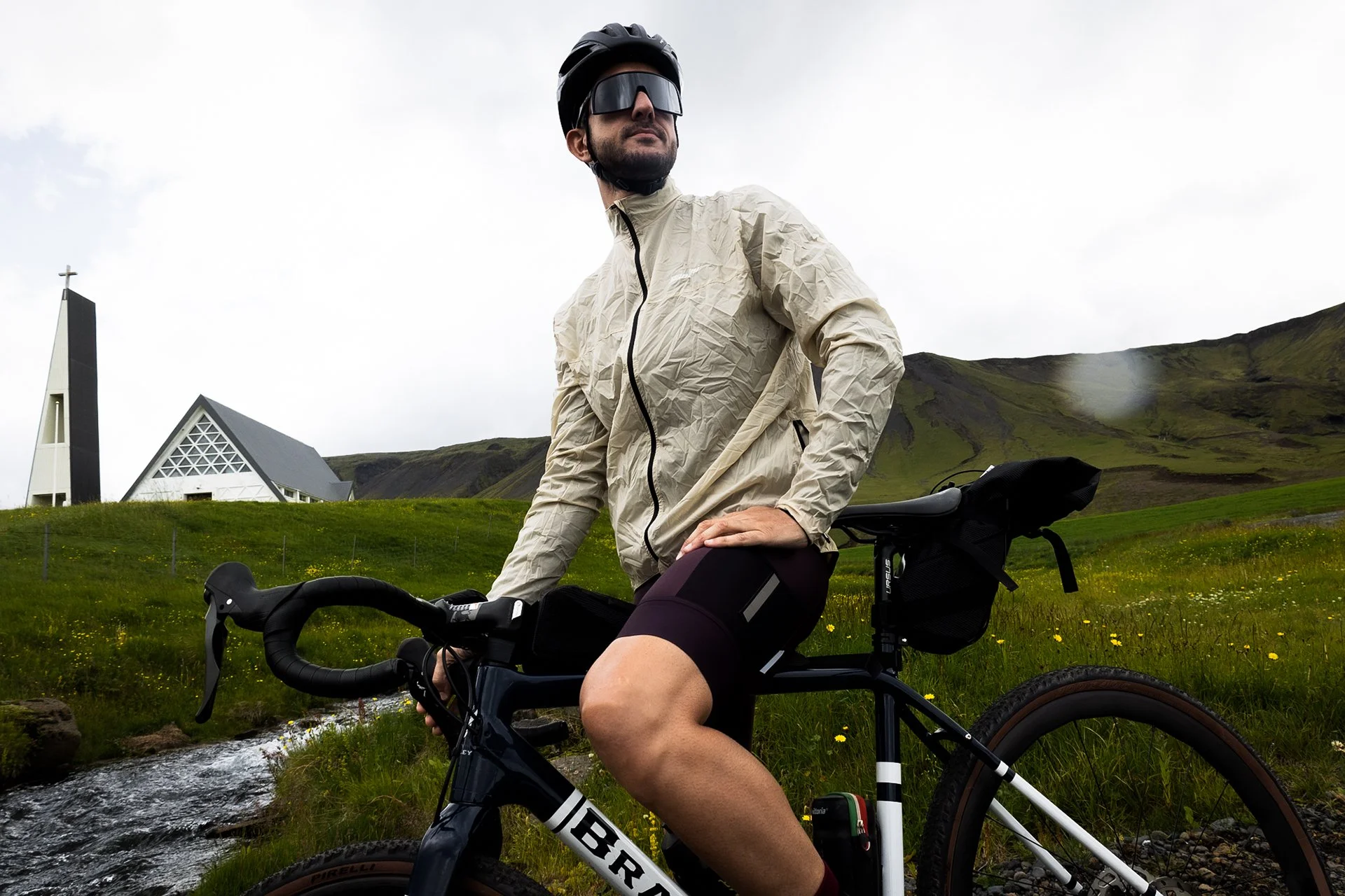 A man in outdoor cycling gear, including a beige jacket, black shorts, and a helmet, sitting on a black and white bicycle in a grassy field with small yellow flowers, near a stream, with a white church and green mountains with patches of snow in the 