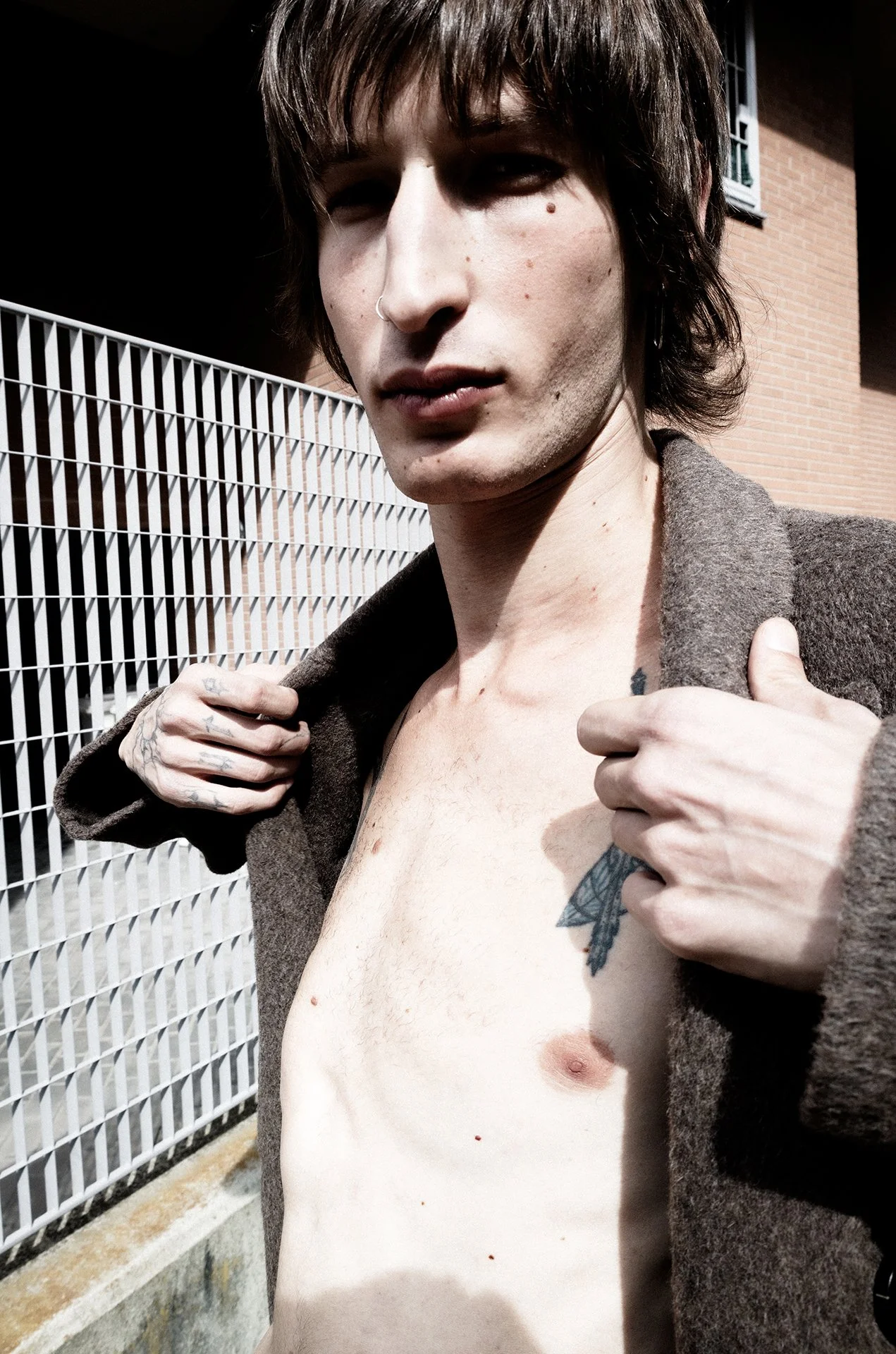 A young person with dark hair, tattoos, and a nose piercing, lifting their open coat to reveal their bare chest, standing outdoors beside a metal fence and a brick building.
