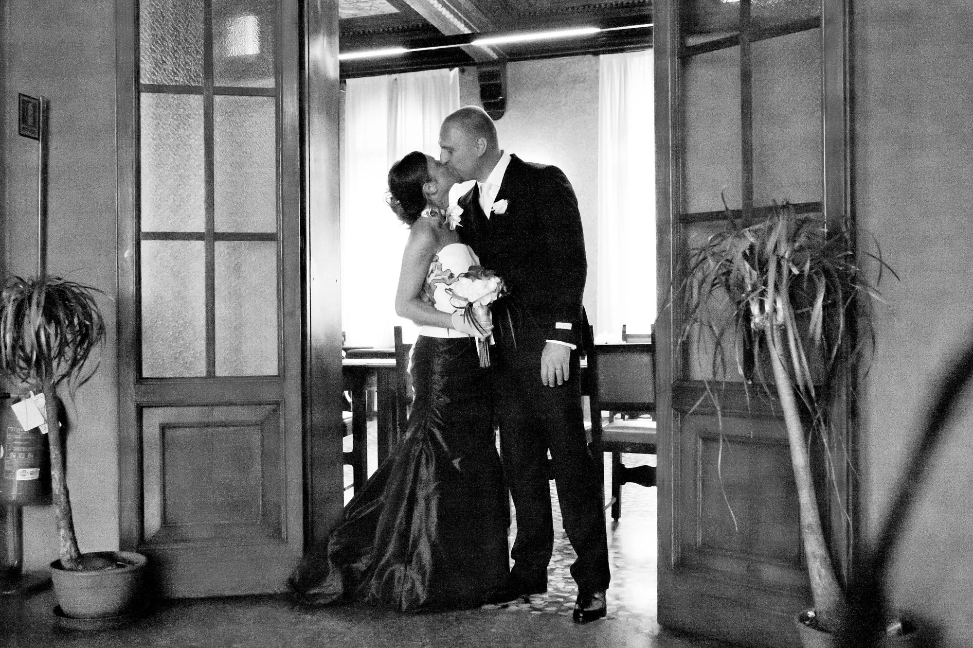 A black-and-white photo of a couple kissing, with the woman holding a bouquet of flowers, in a cozy indoor setting with potted plants and curtains in the background.
