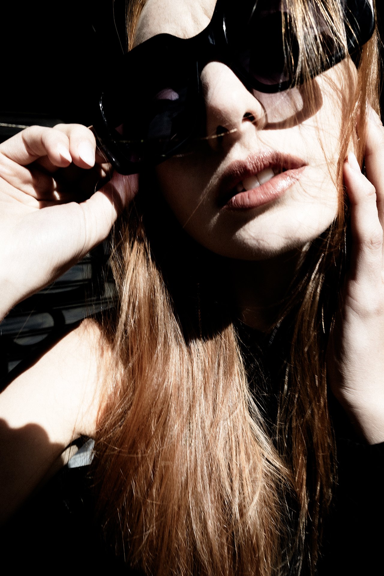 Close-up of a woman with long red hair wearing black sunglasses, touching her face and holding the sunglasses with her right hand.