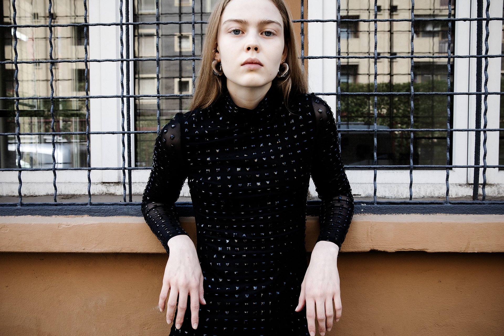 Young woman standing in front of a window with a grid of metal bars, wearing a black dress with small silver details and large hoop earrings.