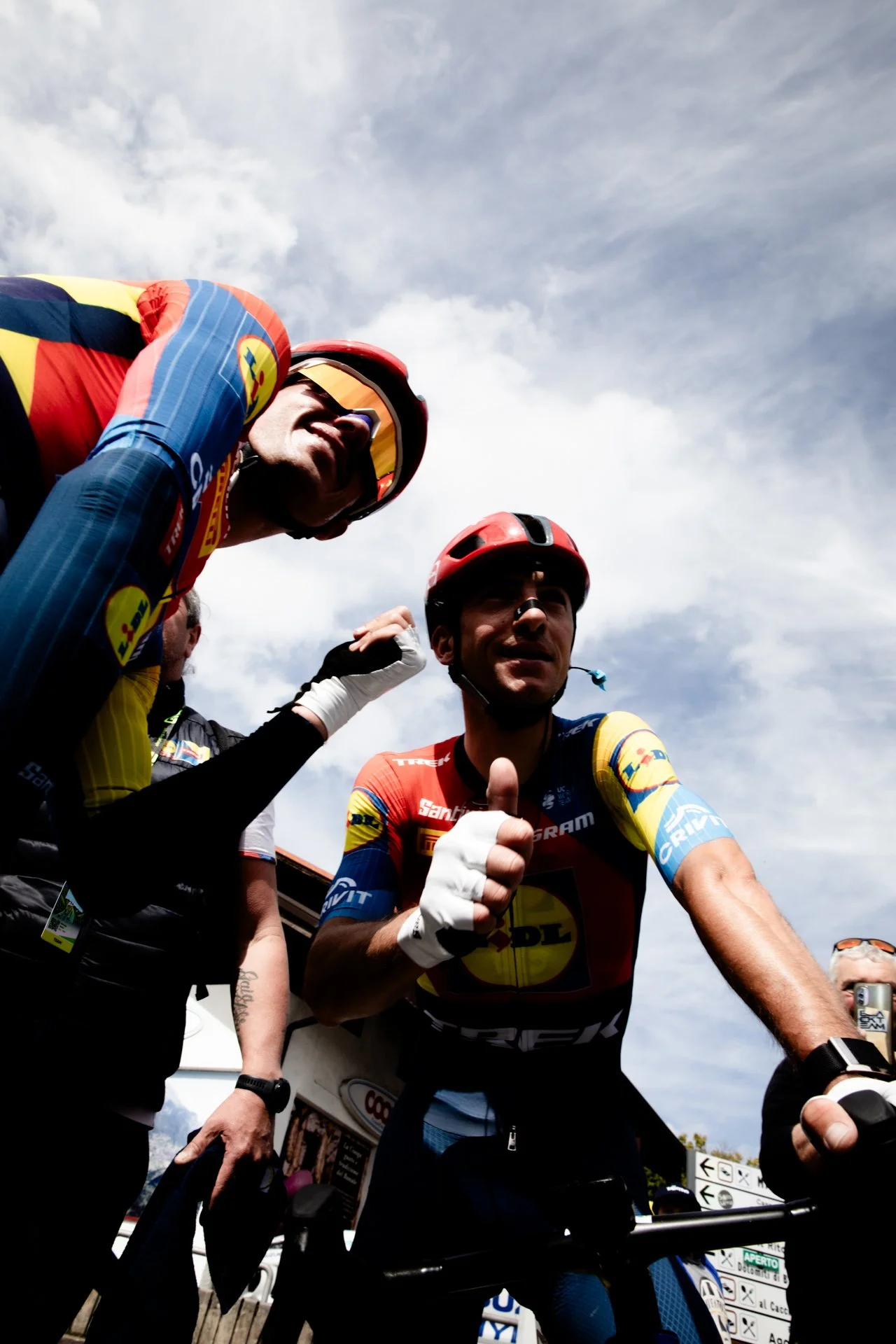 Cyclists in racing gear, helmets, and sunglasses, standing outdoors under a partly cloudy sky, with some giving thumbs-up signs and others adjusting their gear.