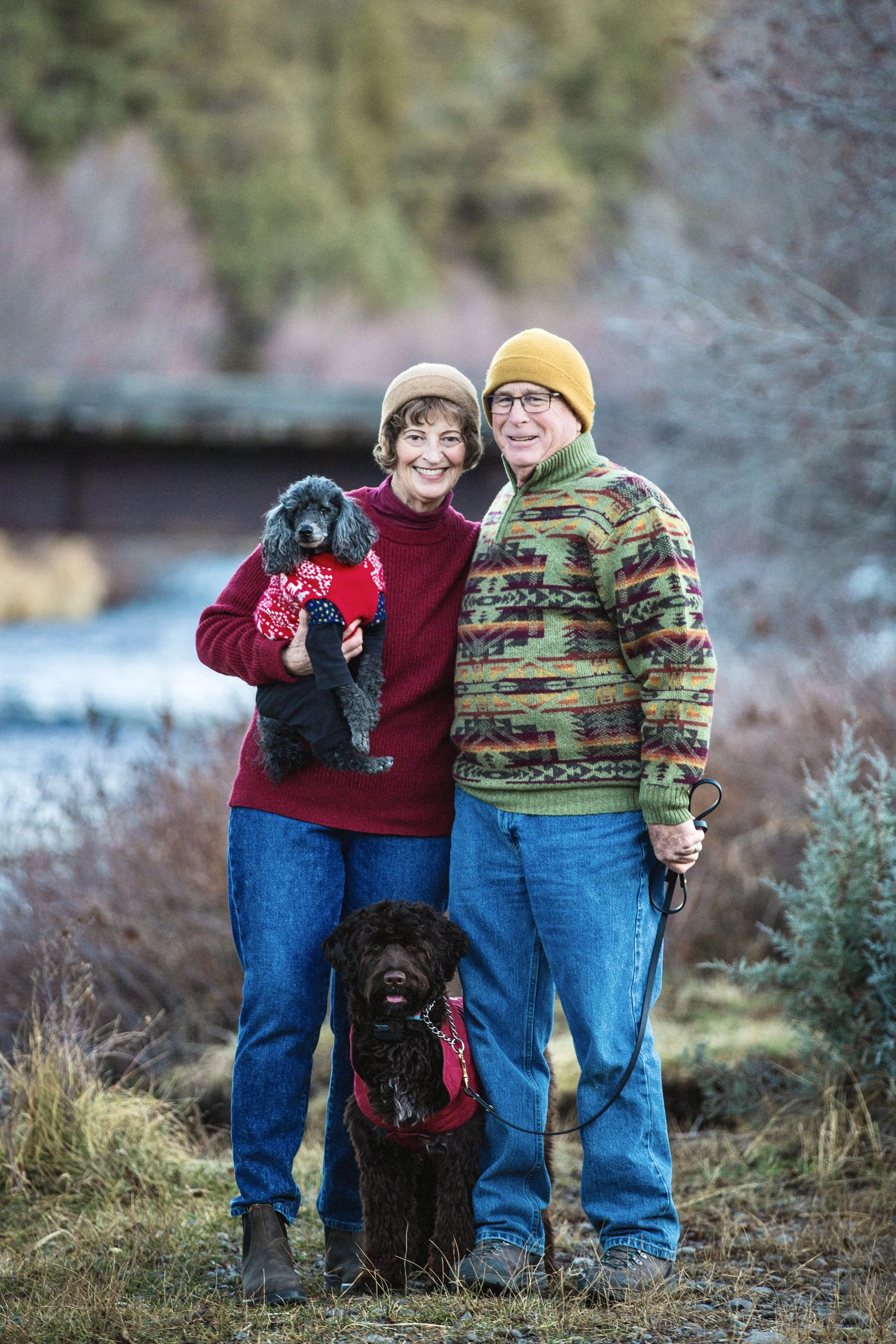 An older couple stands outside in a natural setting with grass and trees, smiling at the camera. The woman is holding a small black poodle wearing a red sweater with a white pattern. The man is holding a leash attached to a larger black dog sitting at their feet, wearing a red vest. Both are dressed warmly in sweaters and beanies.