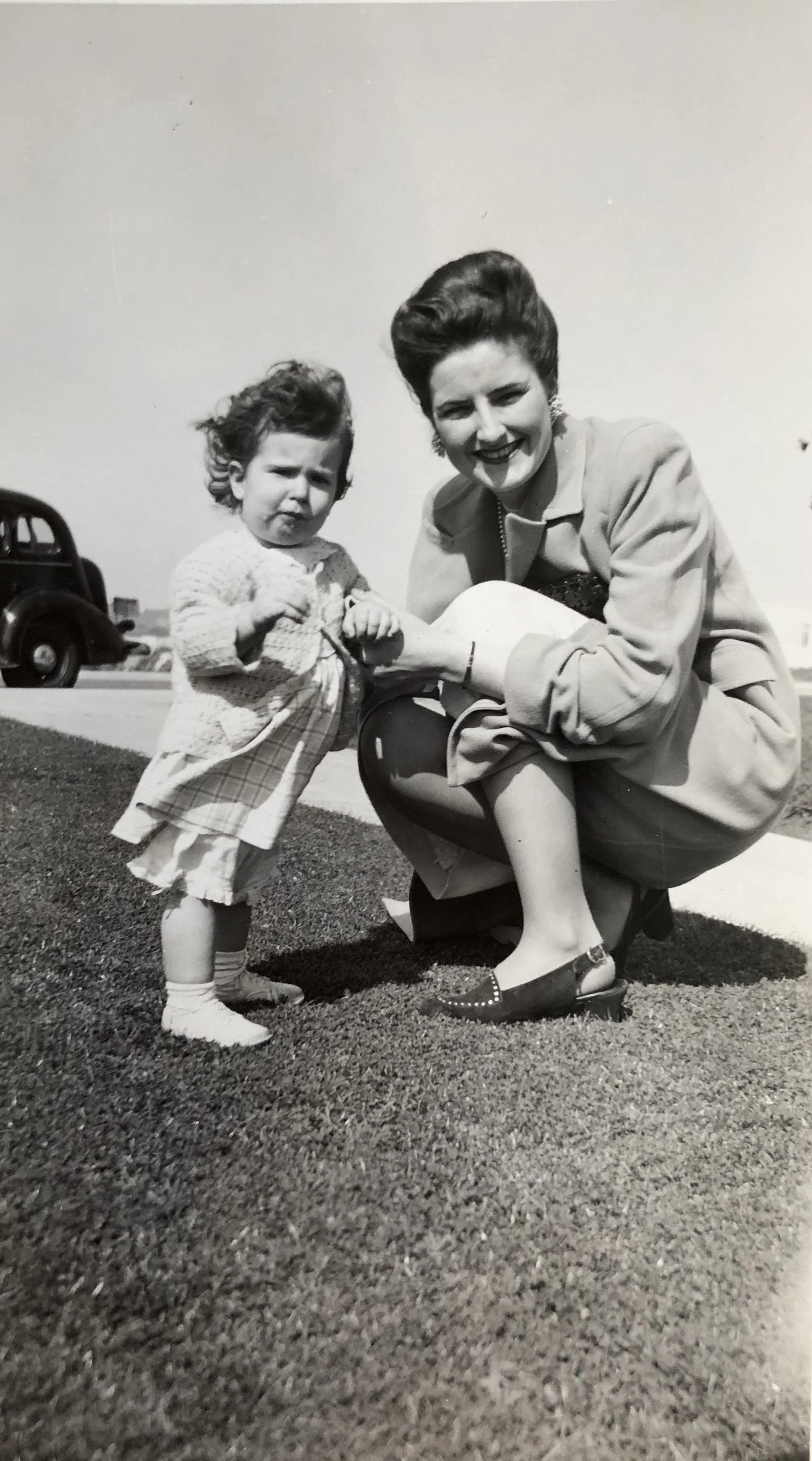 Black and white photo of a young girl and a woman outdoors on grass. The girl is standing with a serious expression, wearing a cardigan, plaid pants, and sneakers. The woman is squatting beside her, smiling, wearing a suit jacket, a skirt, and flats, with her hair styled in an updo.