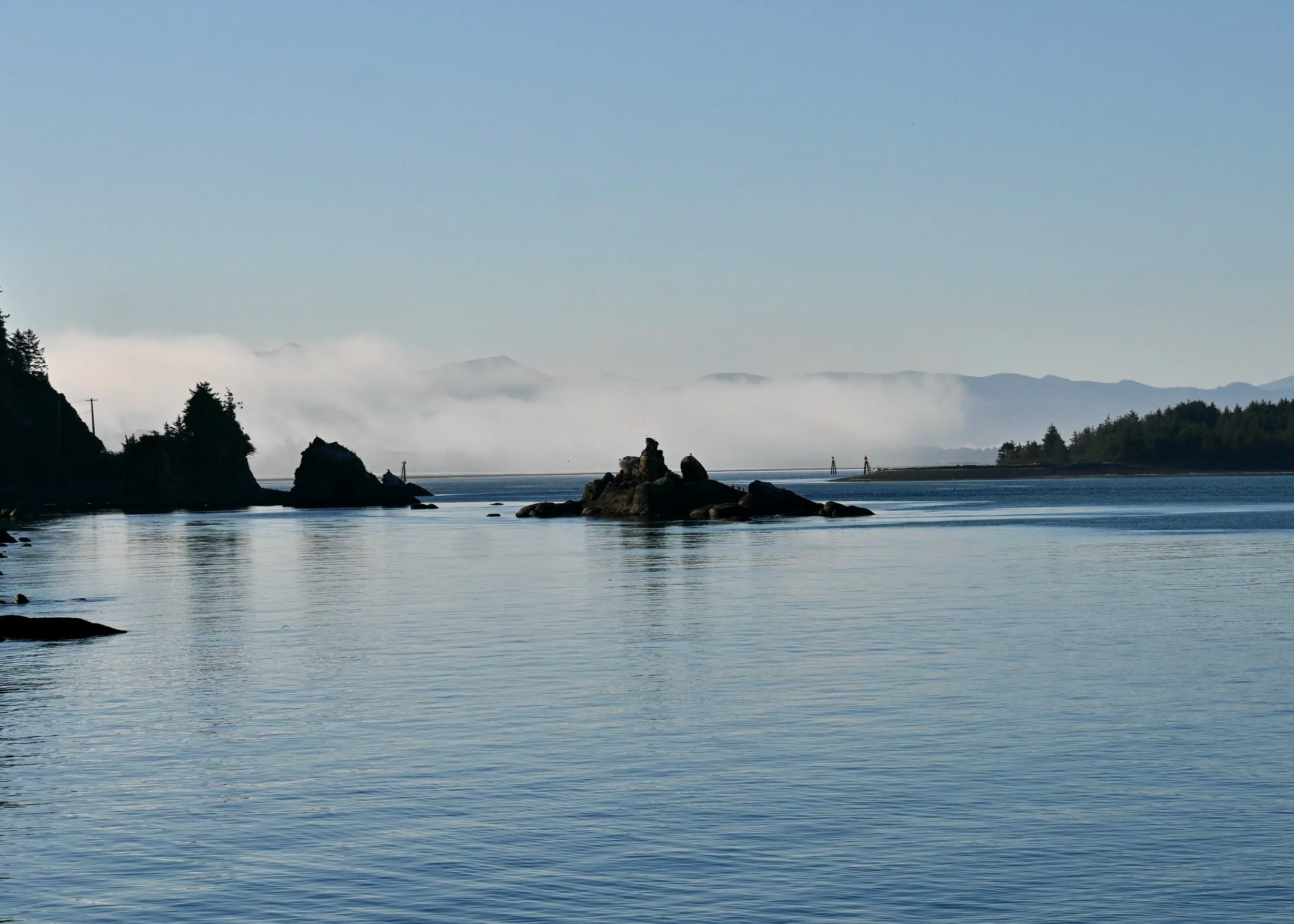 A calm body of water with rocks along the shoreline, surrounded by trees and distant mountains shrouded in fog under a clear blue sky.