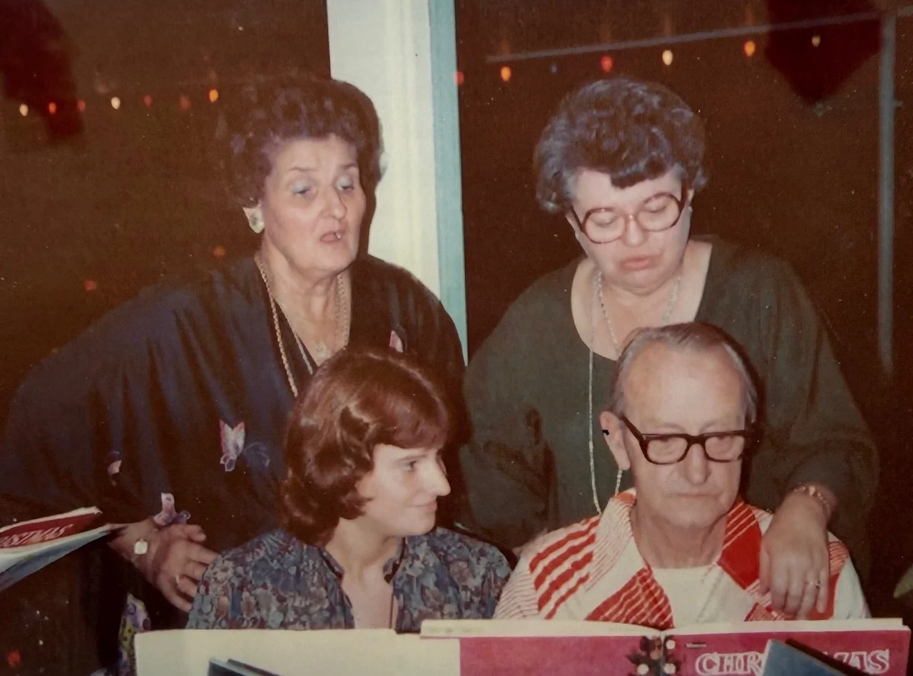 Four people, three women and one man, gathered around a table looking at a menu. The women have gray and reddish hairstyles, one wears glasses, the man has glasses and a white and red vest. It appears to be a social gathering or dinner.