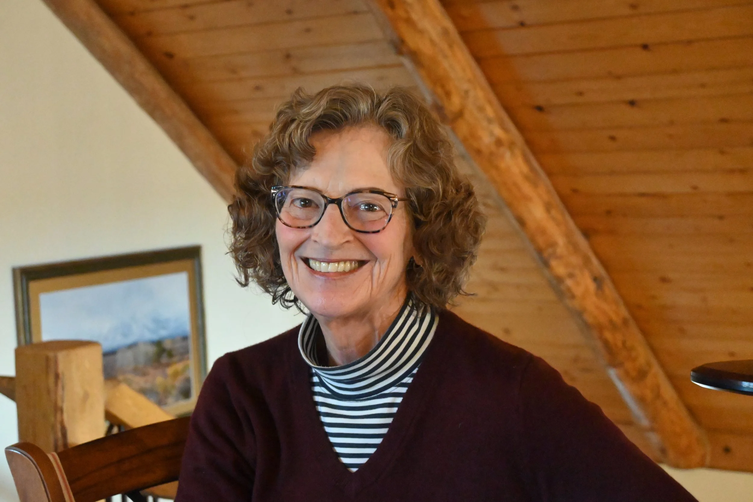 An older woman with curly hair and glasses smiling, sitting indoors with a wooden ceiling and furniture in the background.