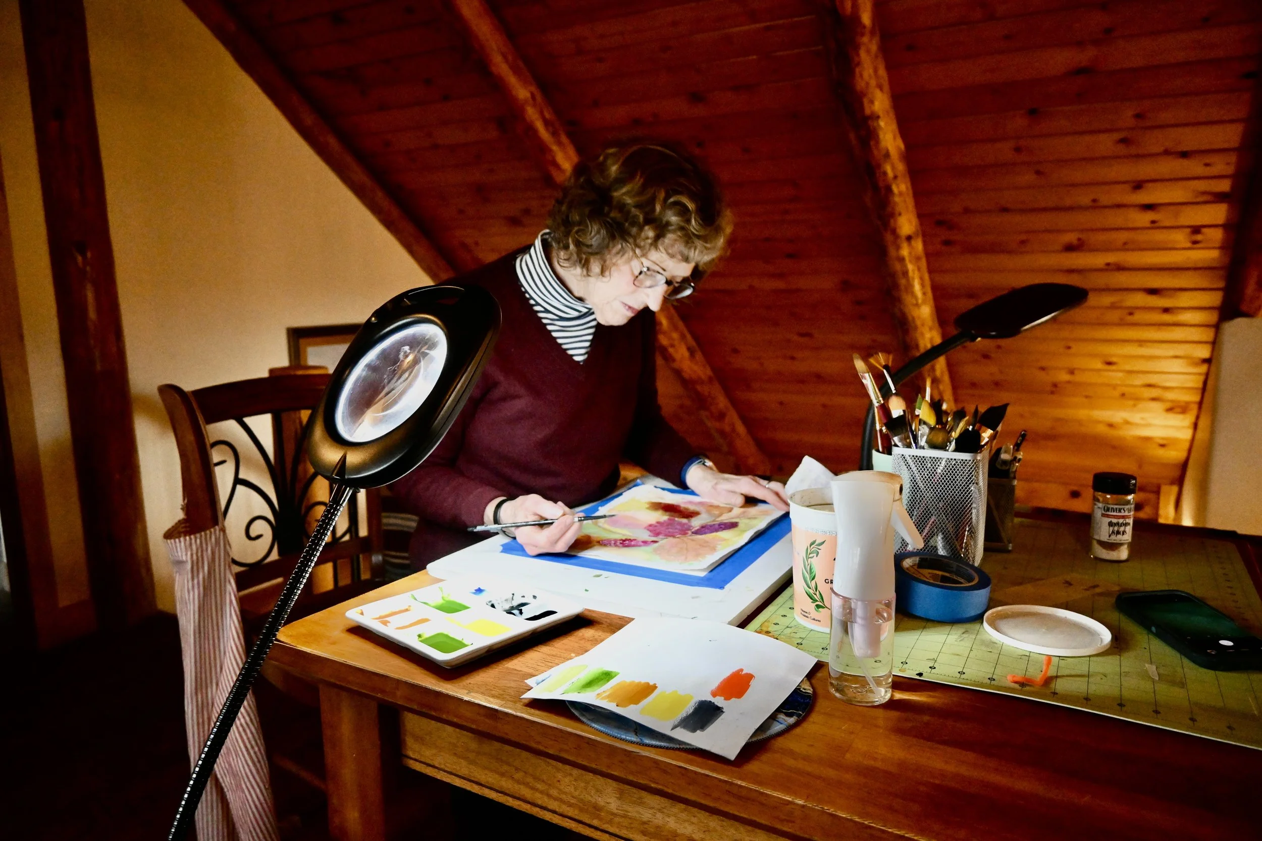 A woman with curly hair wearing glasses, a striped shirt, and a red sweater, sitting at a wooden table in an attic or loft with a sloped wood-paneled ceiling. She is working on an art project with watercolor paints and brushes, surrounded by art supplies including paint palettes, paper, and cups. A lamp is on the table, illuminating her workspace.