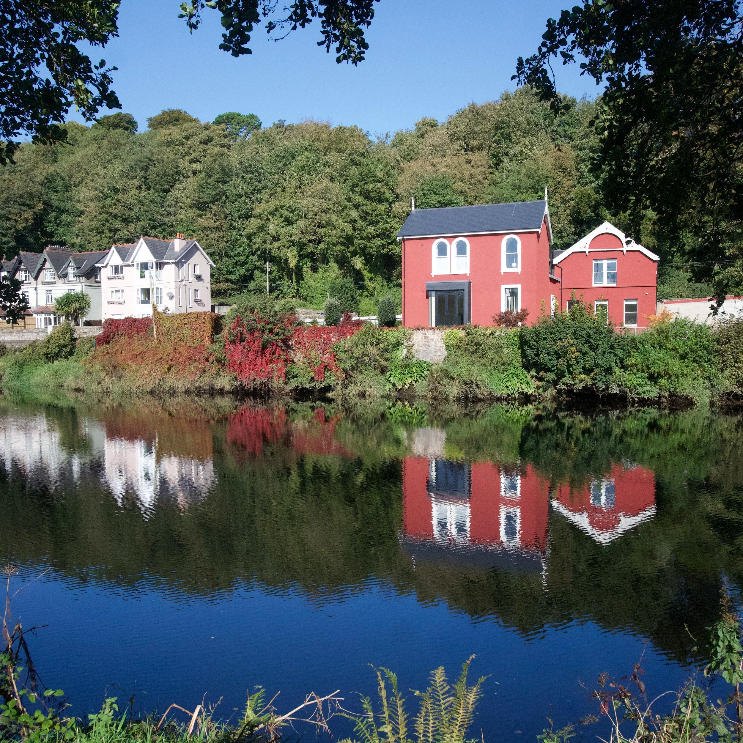 A calm river with reflections of trees and houses along its banks. Houses with varied architectural styles and colors are visible, with a backdrop of a dense green forest under a clear blue sky.