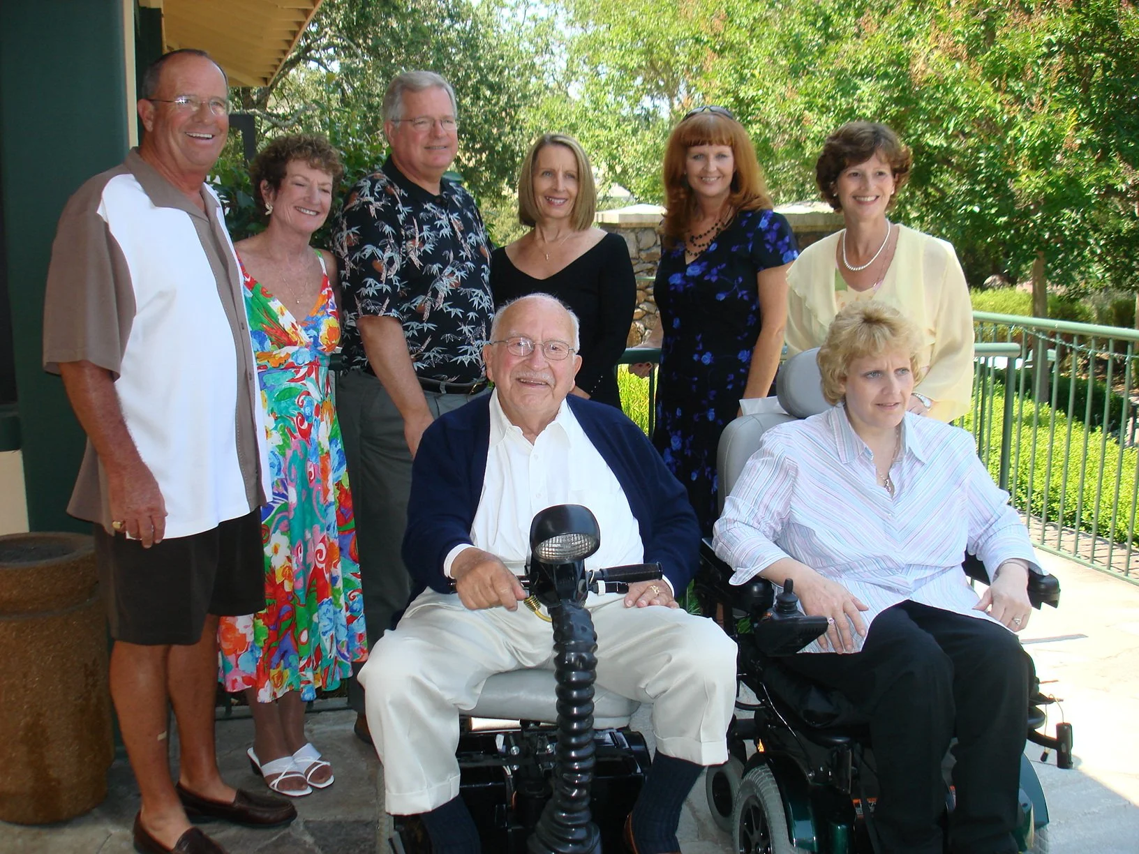 Group of eight people, some in wheelchairs, posing outdoors on a sunny day with green trees in the background.