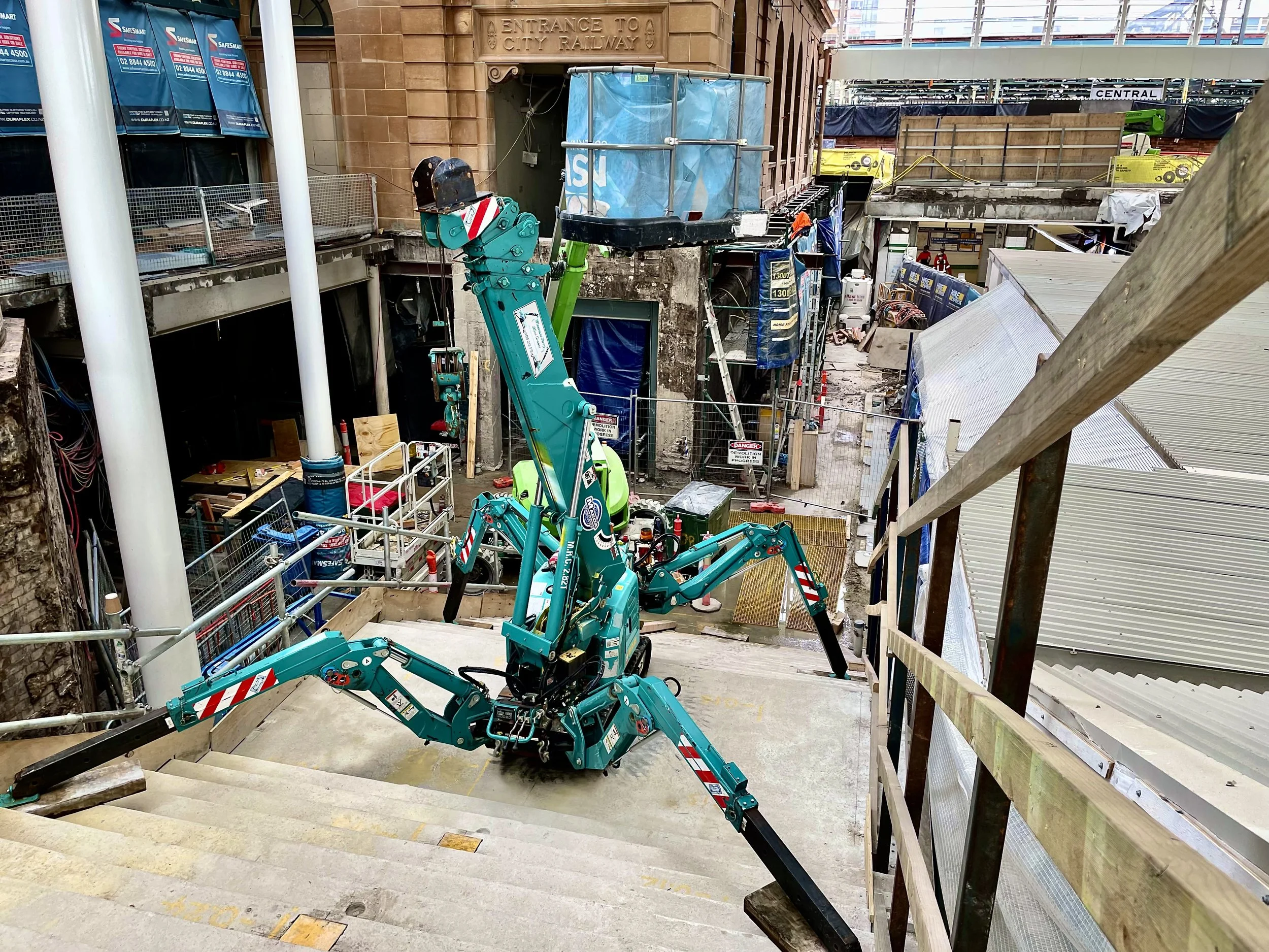 A construction site with a blue spider crane climbing stairs, surrounded by temporary barriers, scaffolding, and construction materials, in an urban area.
