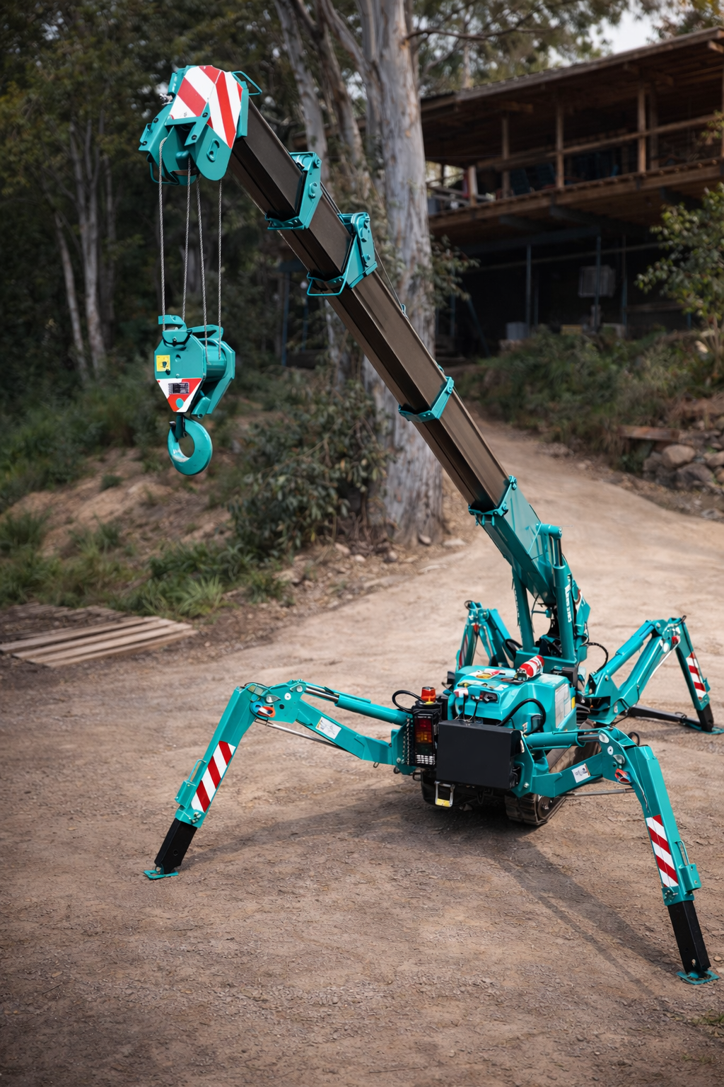 A teal remote-controlled crane with a long adjustable arm and a hook at the end, situated on a dirt path in a wooded area.