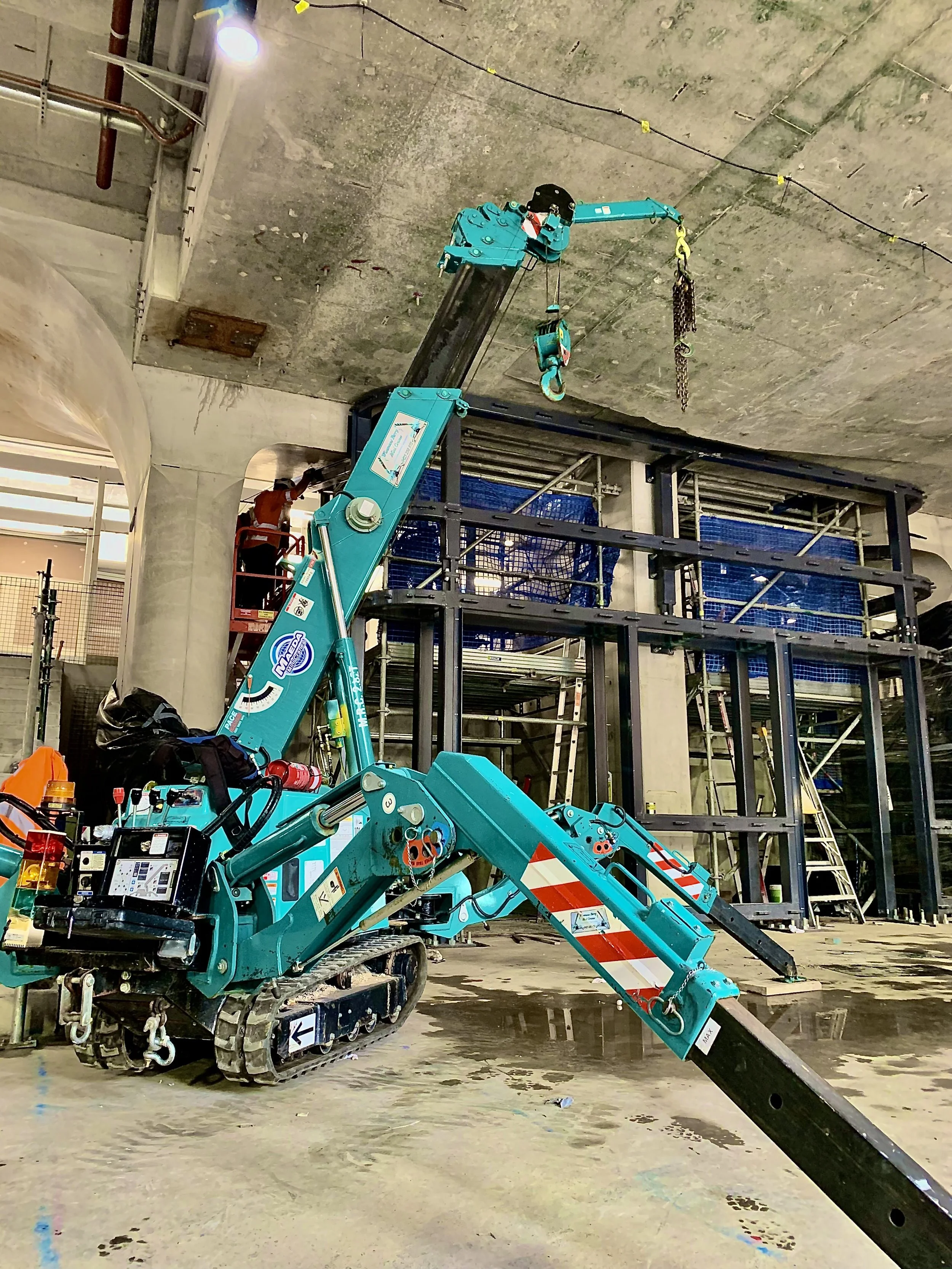 A construction site with a teal mini crane and a worker in the background. The crane is parked on the floor with some water puddles, and there is a steel framework for a building structure behind it.