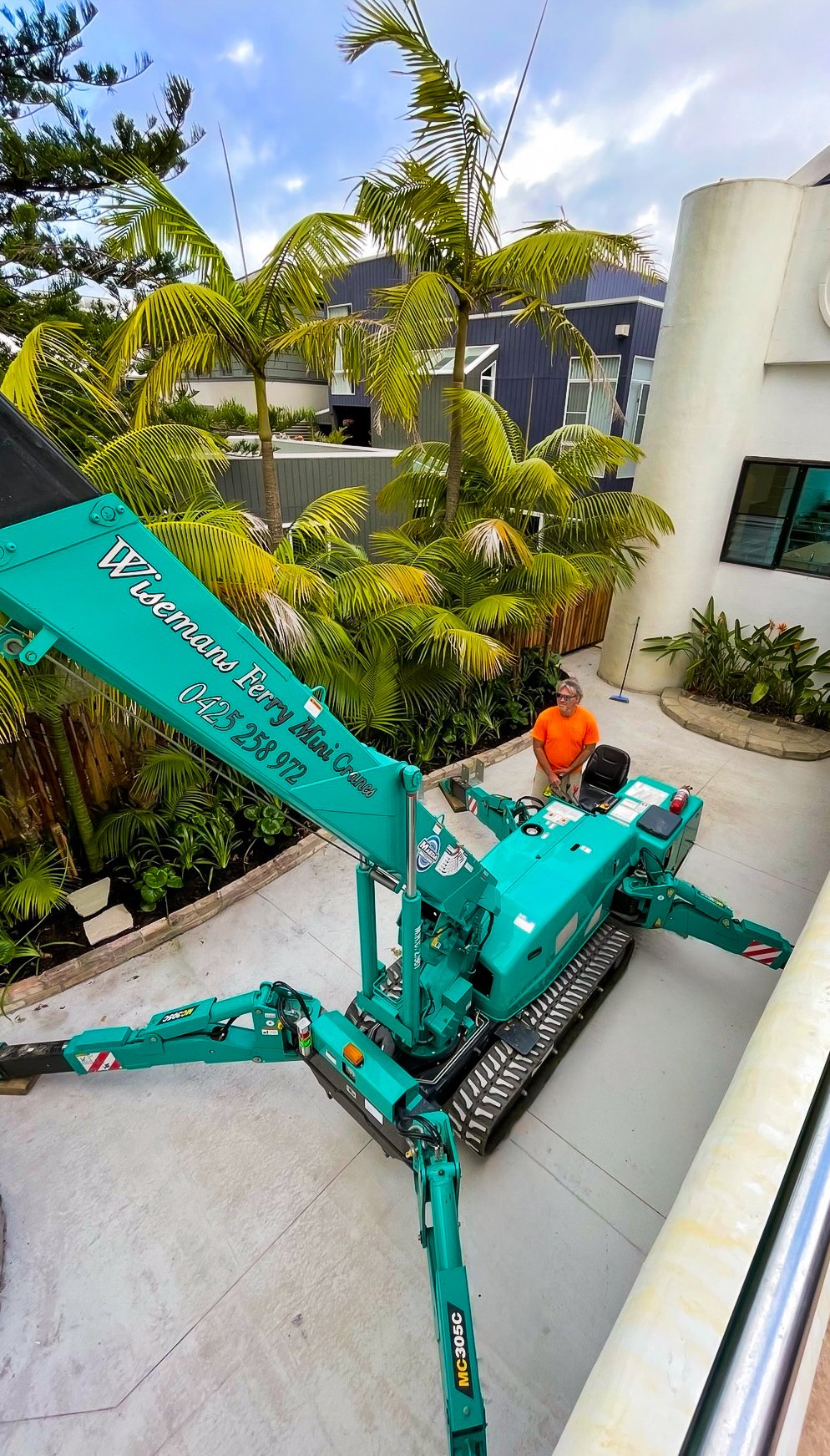 A man in an orange shirt stands near a teal-colored crane parked on a concrete surface in a residential area with palm trees and modern houses in the background.