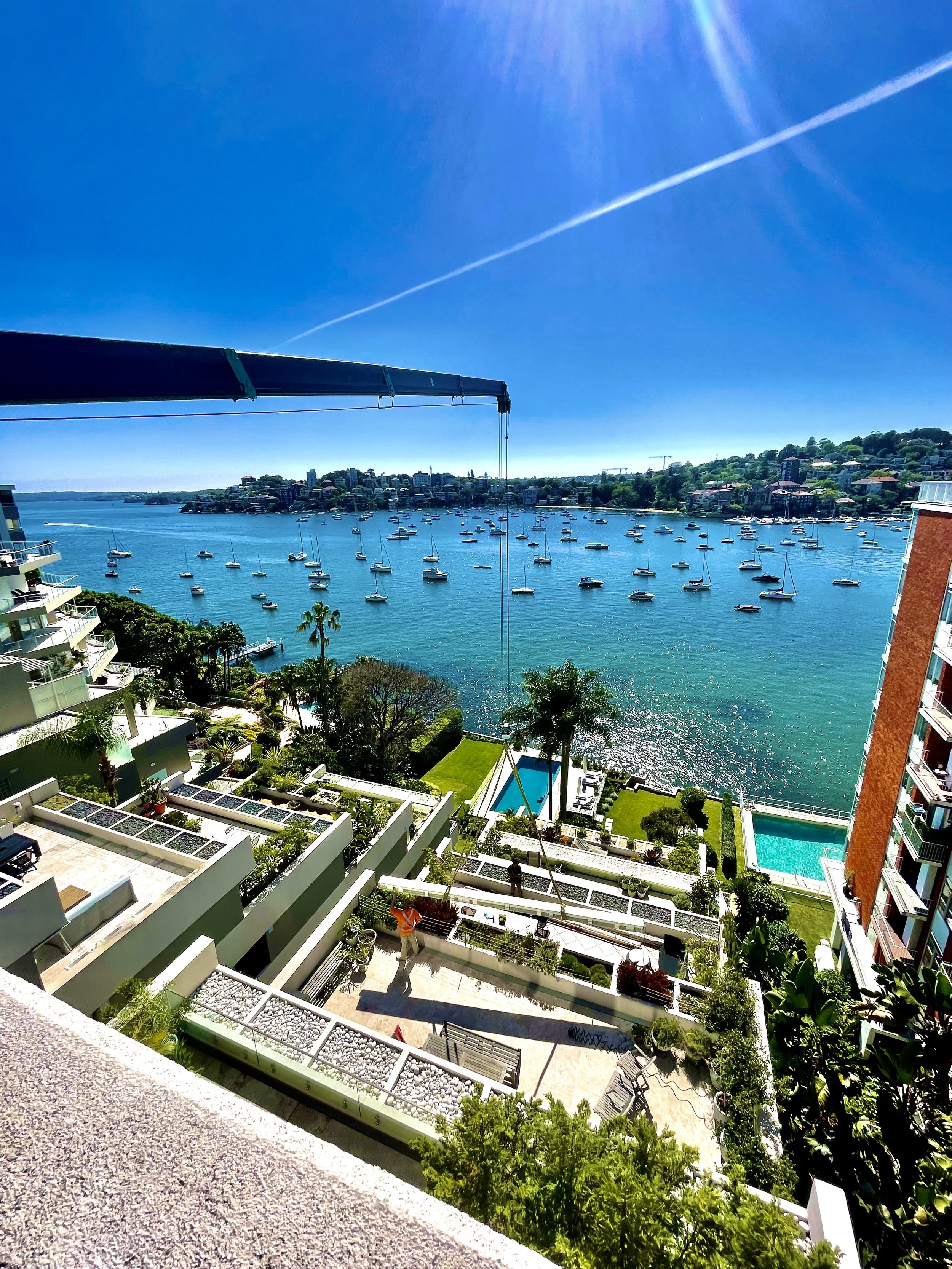View of a bay with numerous sailboats, a clear blue sky with a contrail, and part of a balcony and garden area of a high-rise building.