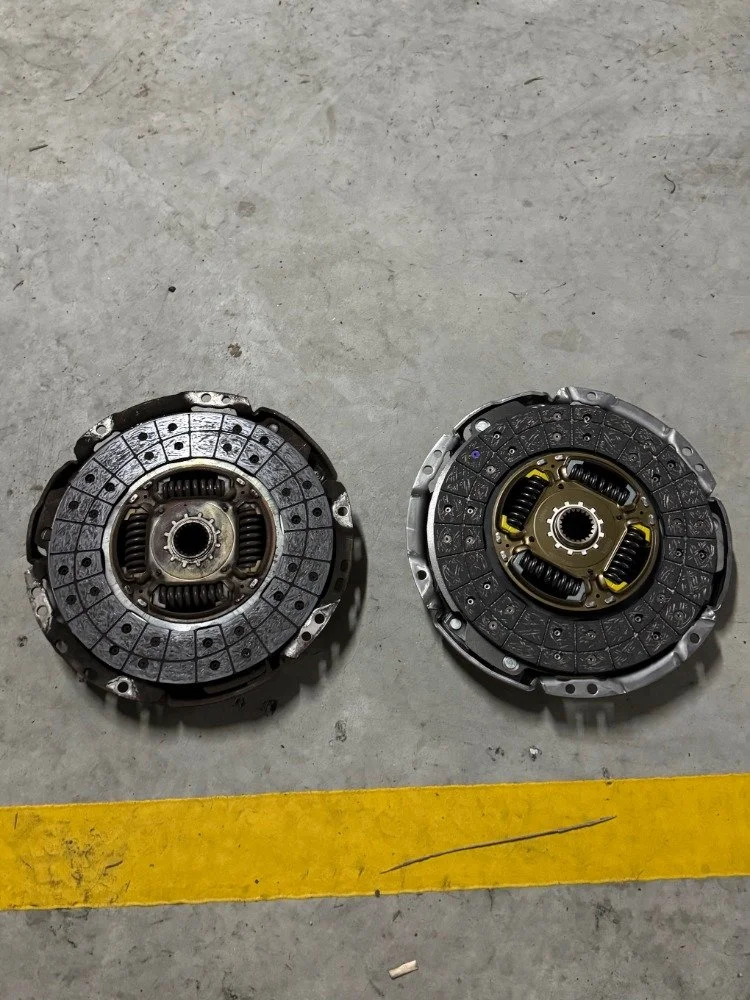 Two automotive clutch pressure plates placed on a concrete floor next to a yellow line.