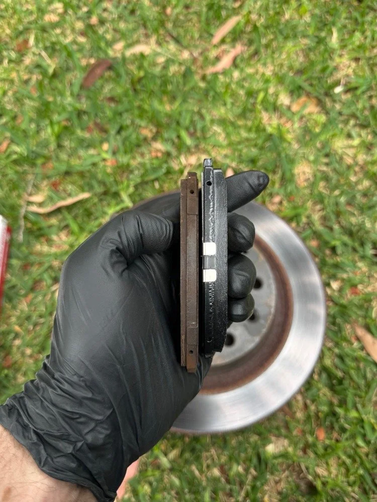 Person wearing a black glove holding a brake pad in front of a car's brake disc on grass.