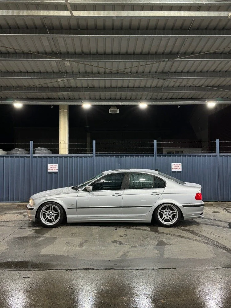 Silver sedan car parked in a wet parking lot at night, behind a blue metal fence with caution signs and a covered area overhead.