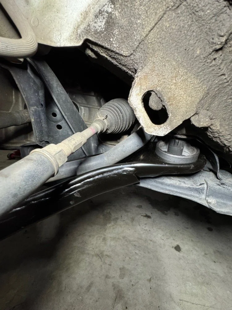 Under the vehicle, showing a damaged or missing fan mount bracket near the oil pan, with various suspension and steering components visible.