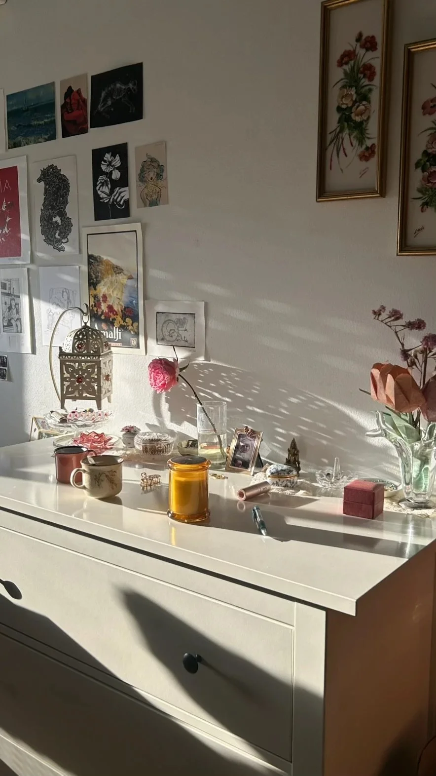 A white dresser with various small decorative items, including a glass vase with pink flowers, candles, a framed photograph, and jewelry. Above the dresser, a wall displays art prints and framed paintings of flowers. Sunlight casts shadows across the scene.