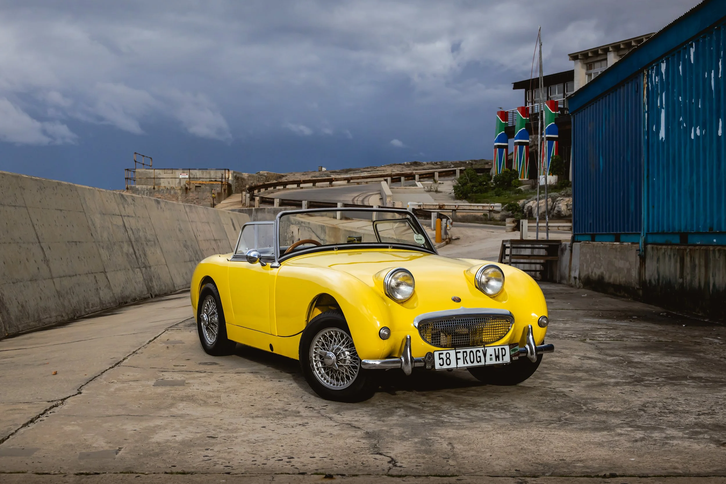 Yellow vintage convertible sports car parked on a concrete surface, with stormy clouds overhead and a blue building on the right.