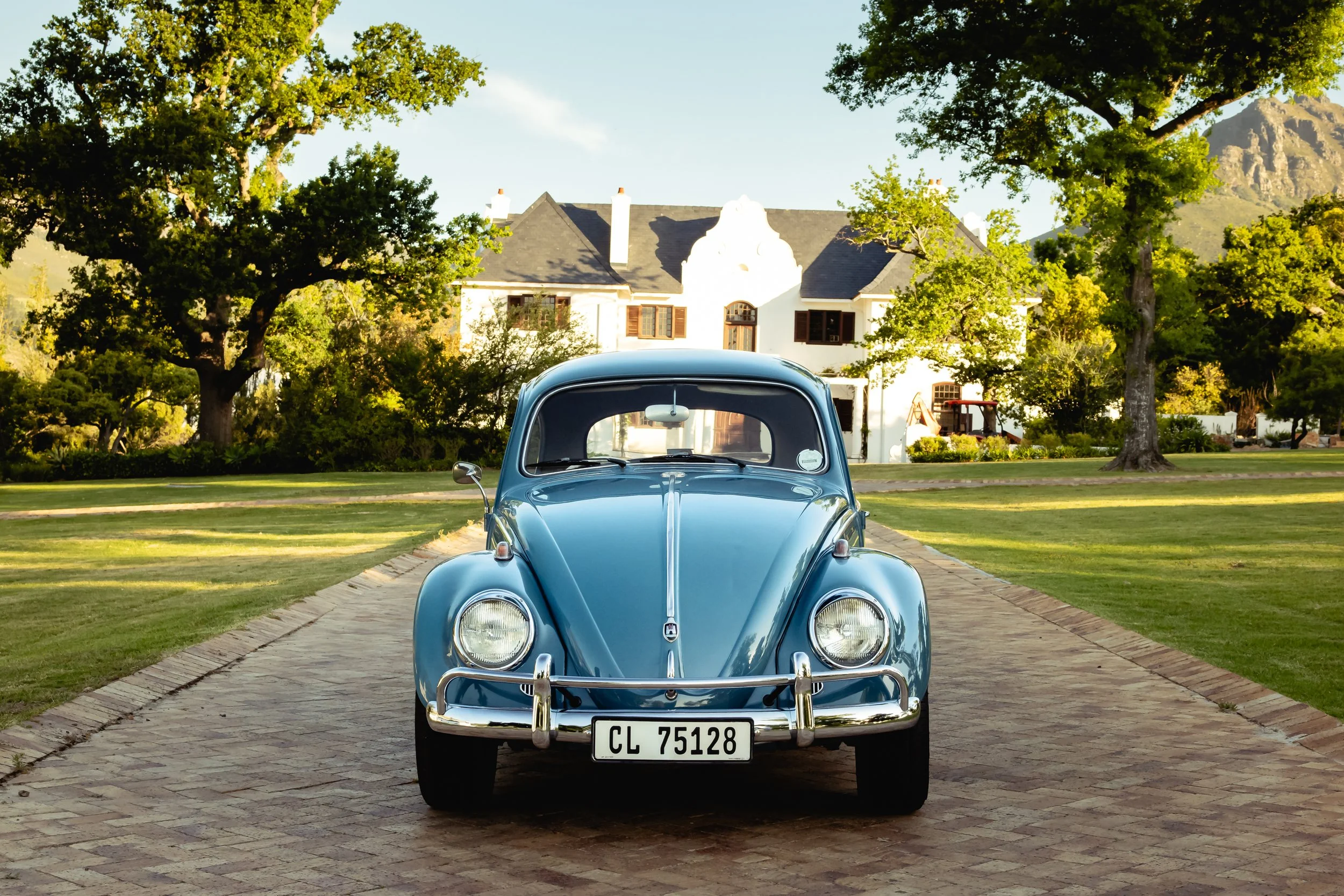 A vintage blue Volkswagen Beetle parked on a brick driveway in front of a white house with brown shutters, surrounded by green trees and a lawn.
