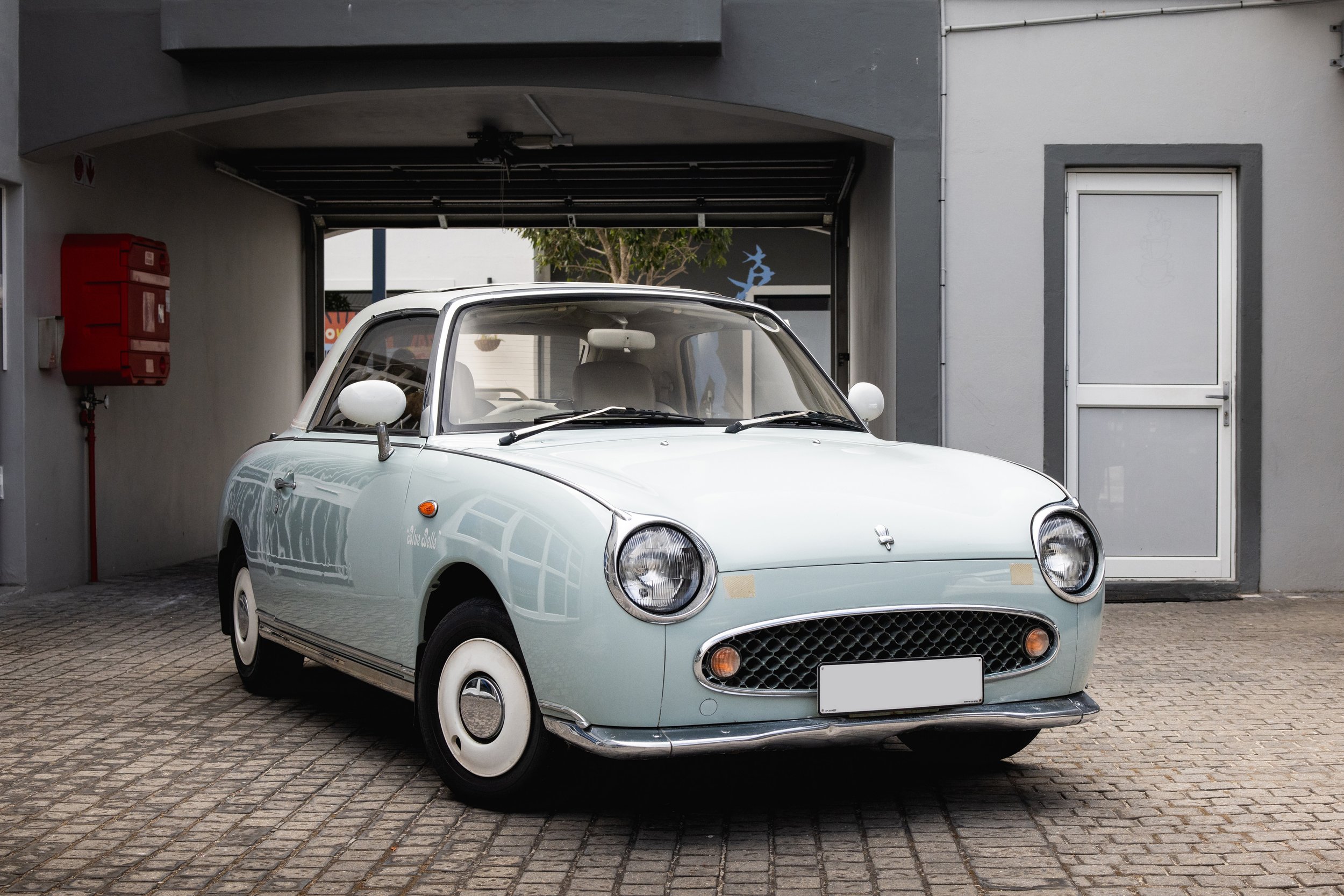 Light blue Nissan Figaro vintage compact car with round headlights parked in front of a gray building, with a garage door and red fire alarm box nearby.