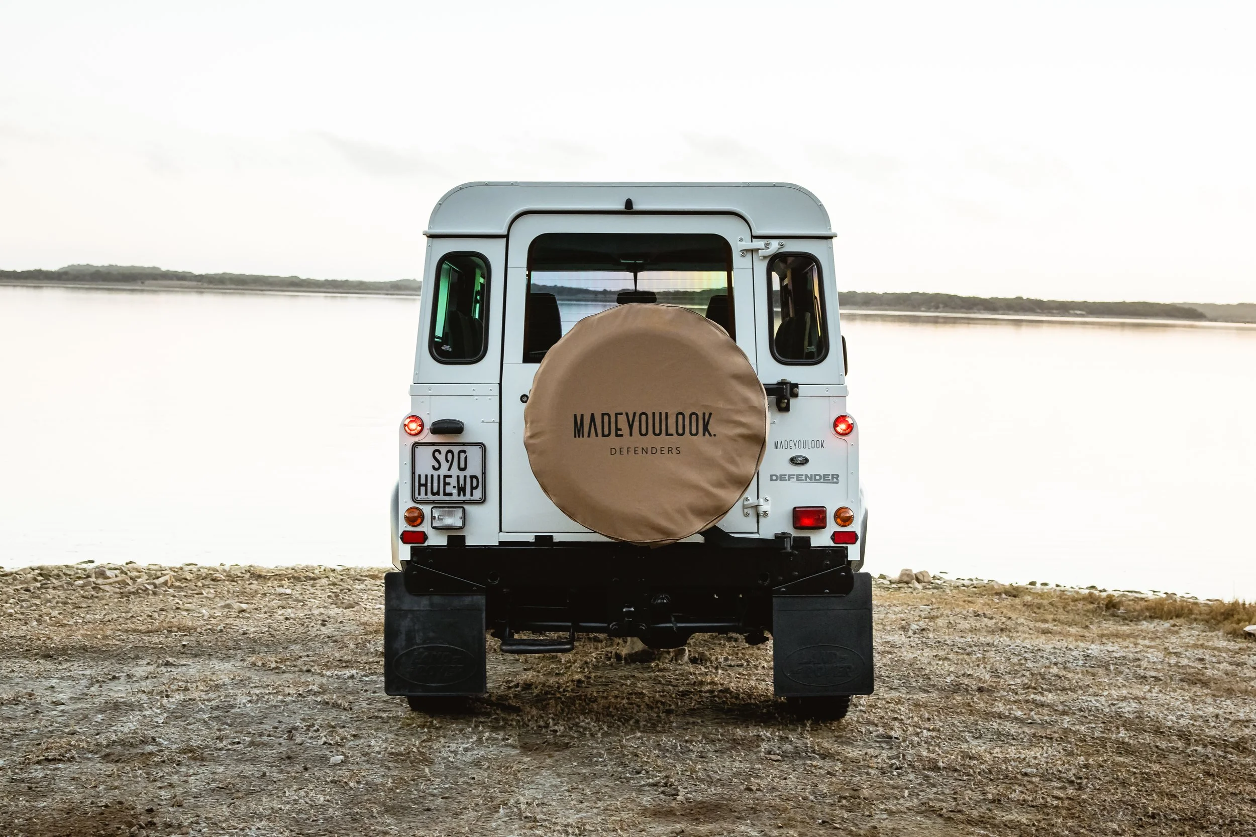 White Land Rover Defender 90 off-road vehicle parked on dirt near a body of water with a view of the horizon in the background.
