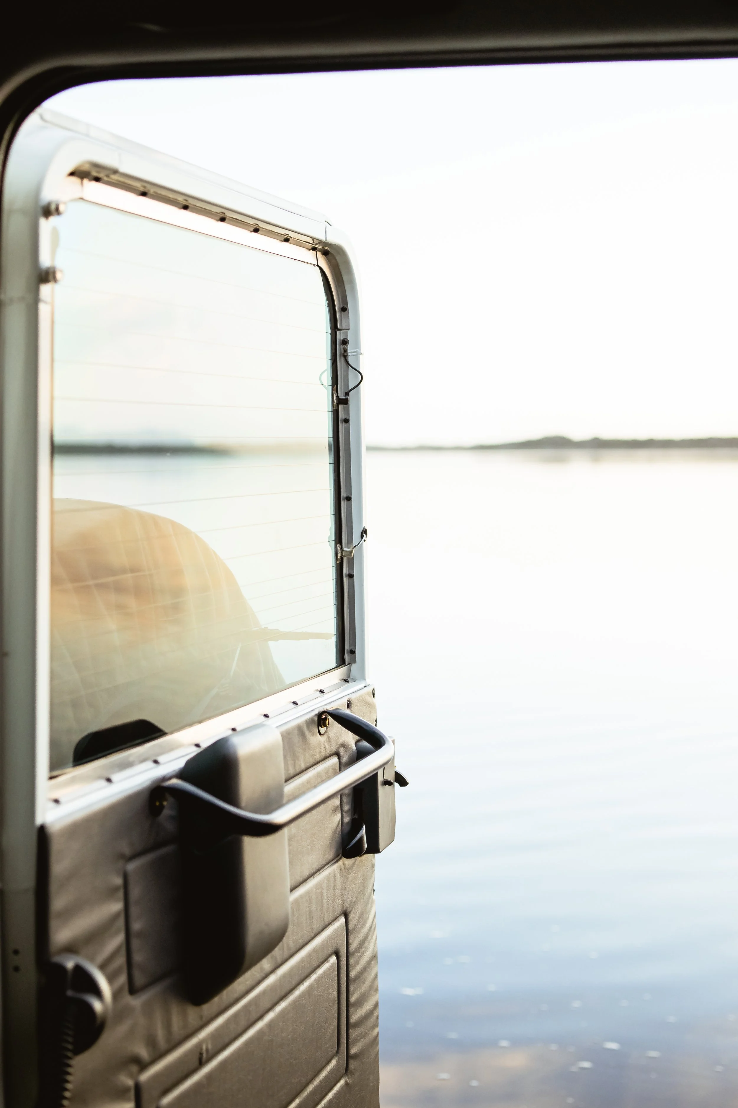 View from inside a Land Rover off-road vehicle looking out at a calm lake or river with a clear sky, with part of the vehicle's rear window and door visible in the foreground.