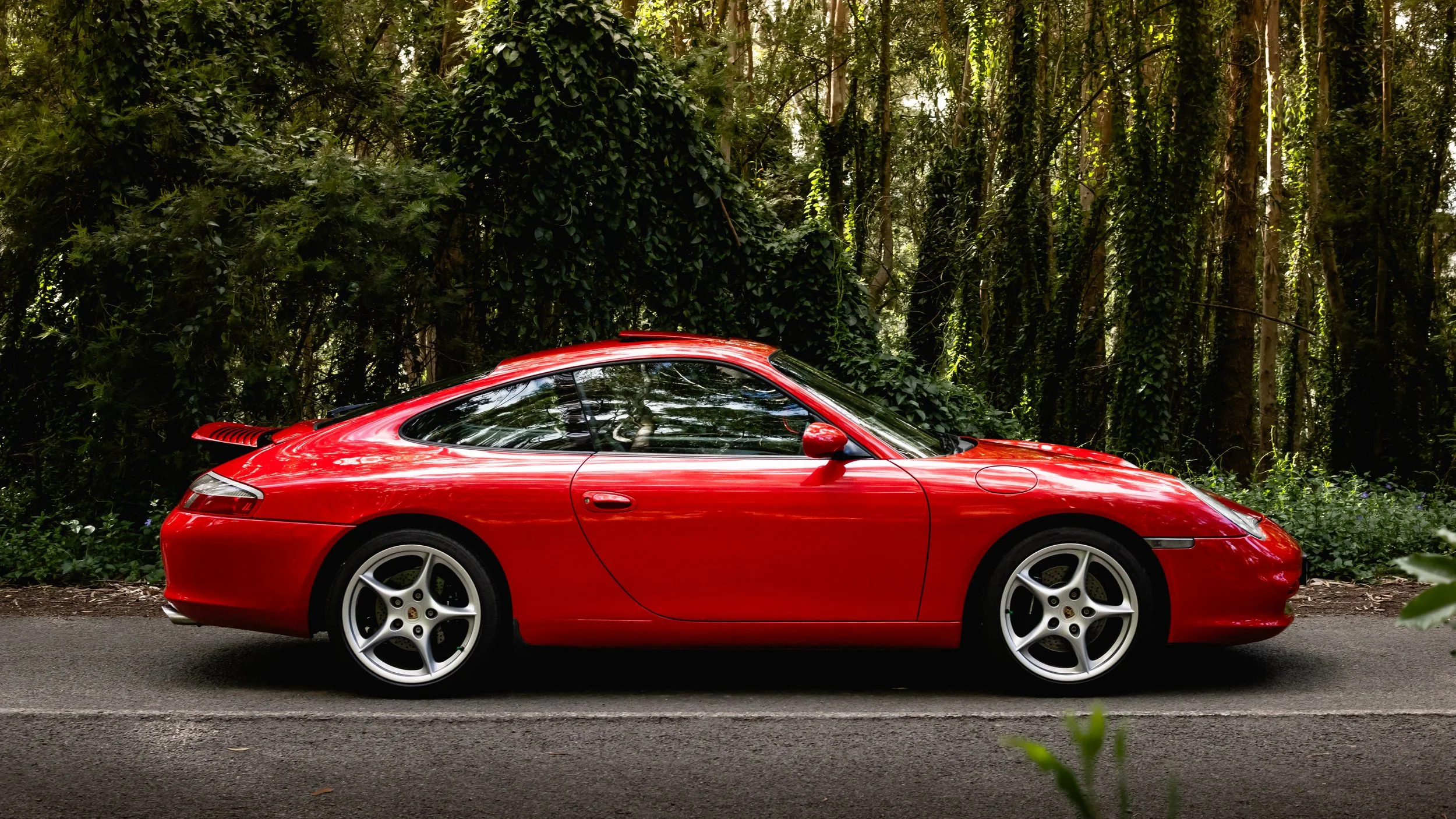 A red Porsche 996 sports car parked on a rural road next to a dense forest with green foliage and tall trees.