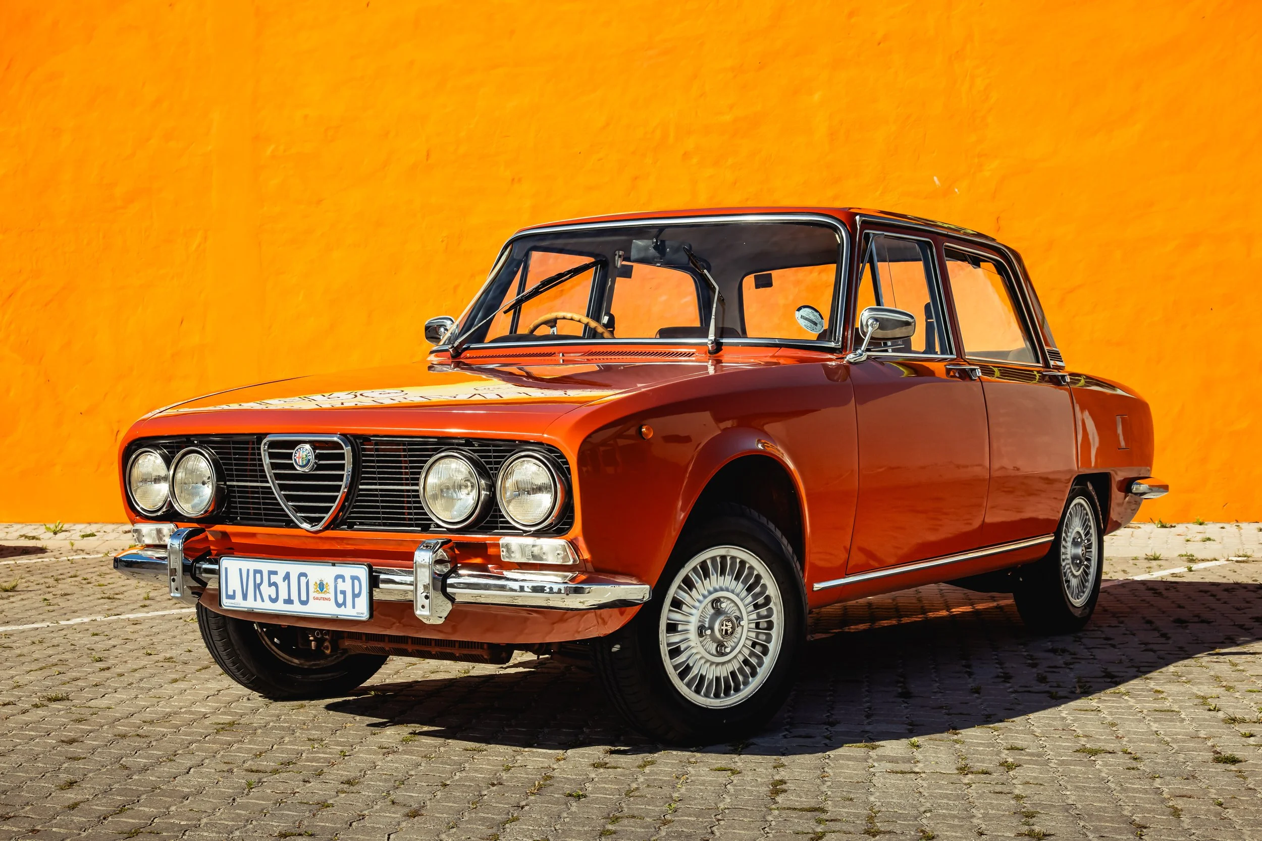 A vintage orange Alfa Romeo Berlina sportscar parked on a brick pavement in front of an orange wall.