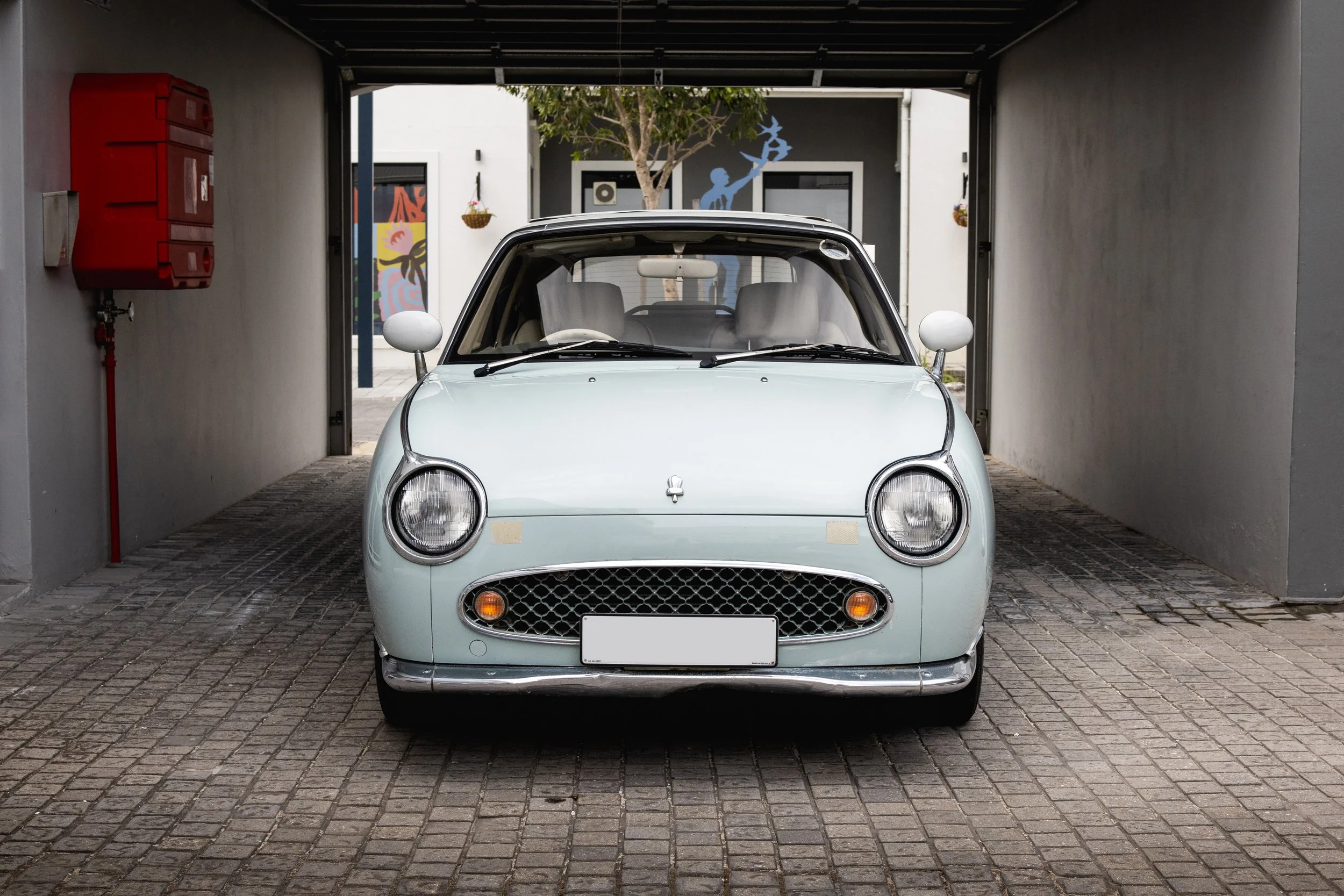 A vintage light blue Nissan Figaro compact car parked in a driveway, viewed from the front.