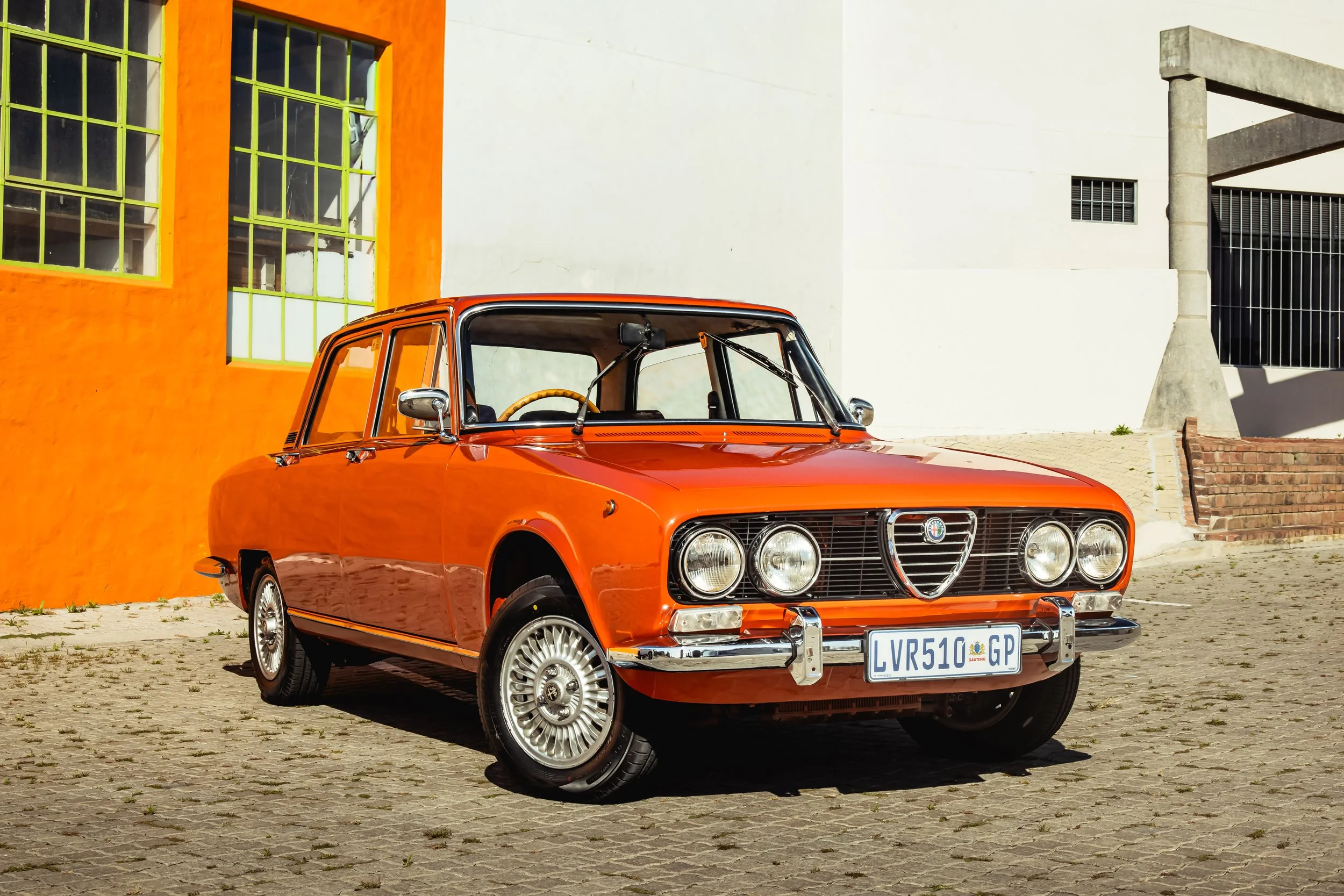 An orange vintage Alfa Romeo car parked on a cobblestone street, with a colorful building with large windows in the background.