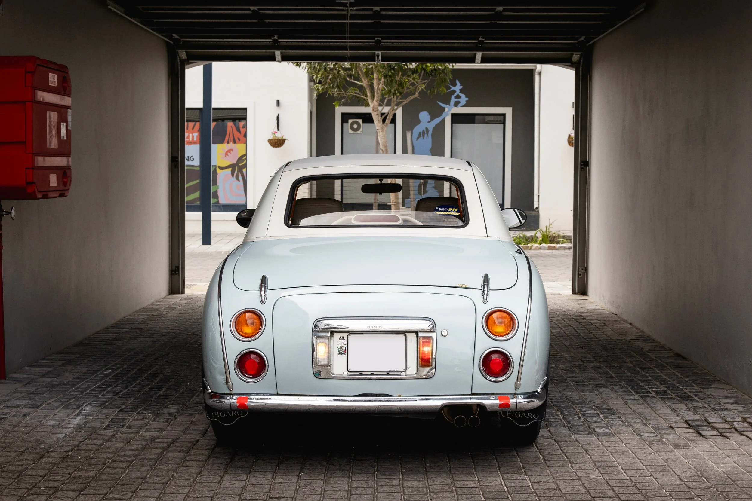 A vintage white car parked in a covered parking space, facing outward, with a tree and modern building in the background.
