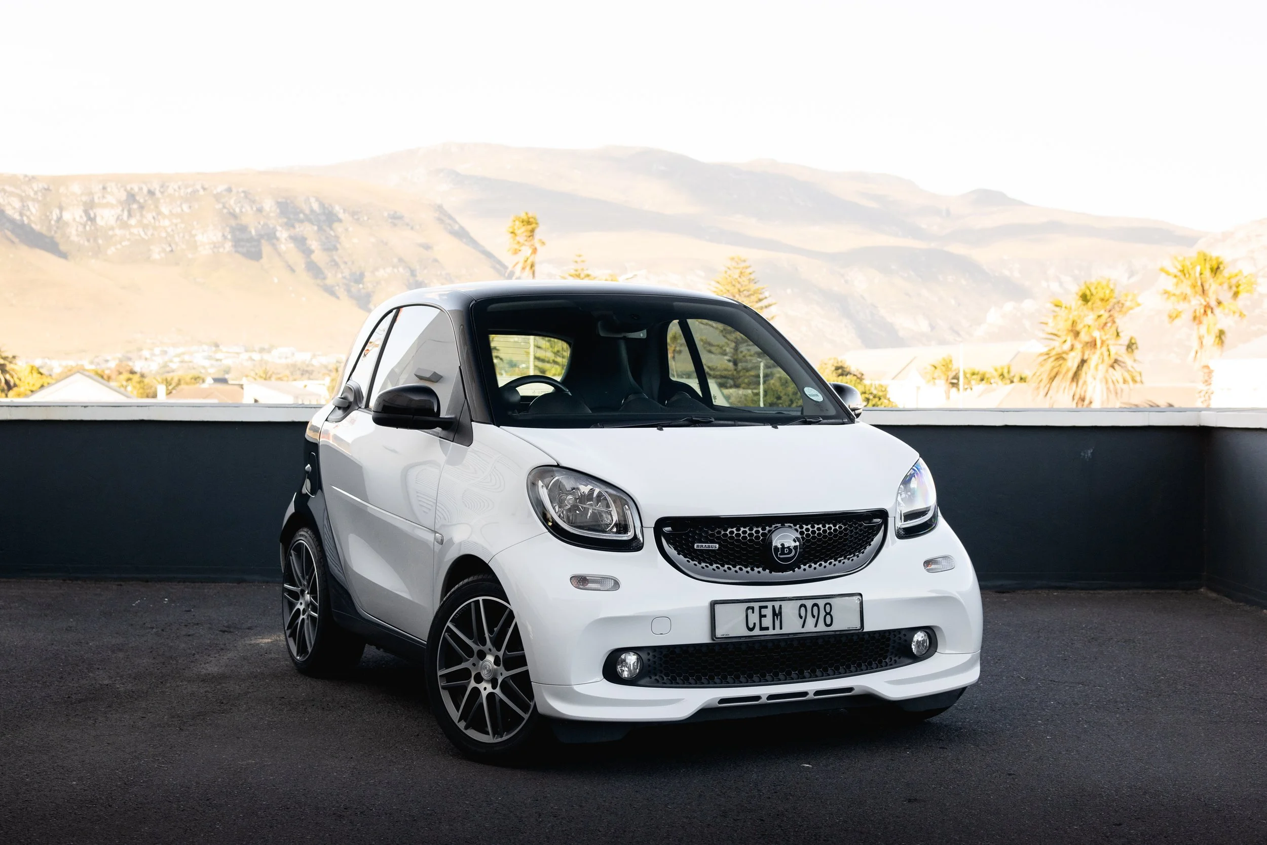 A white Brabus Smart car with custom black wheels parked on an outdoor lot with mountainous landscape and palm trees in the background.