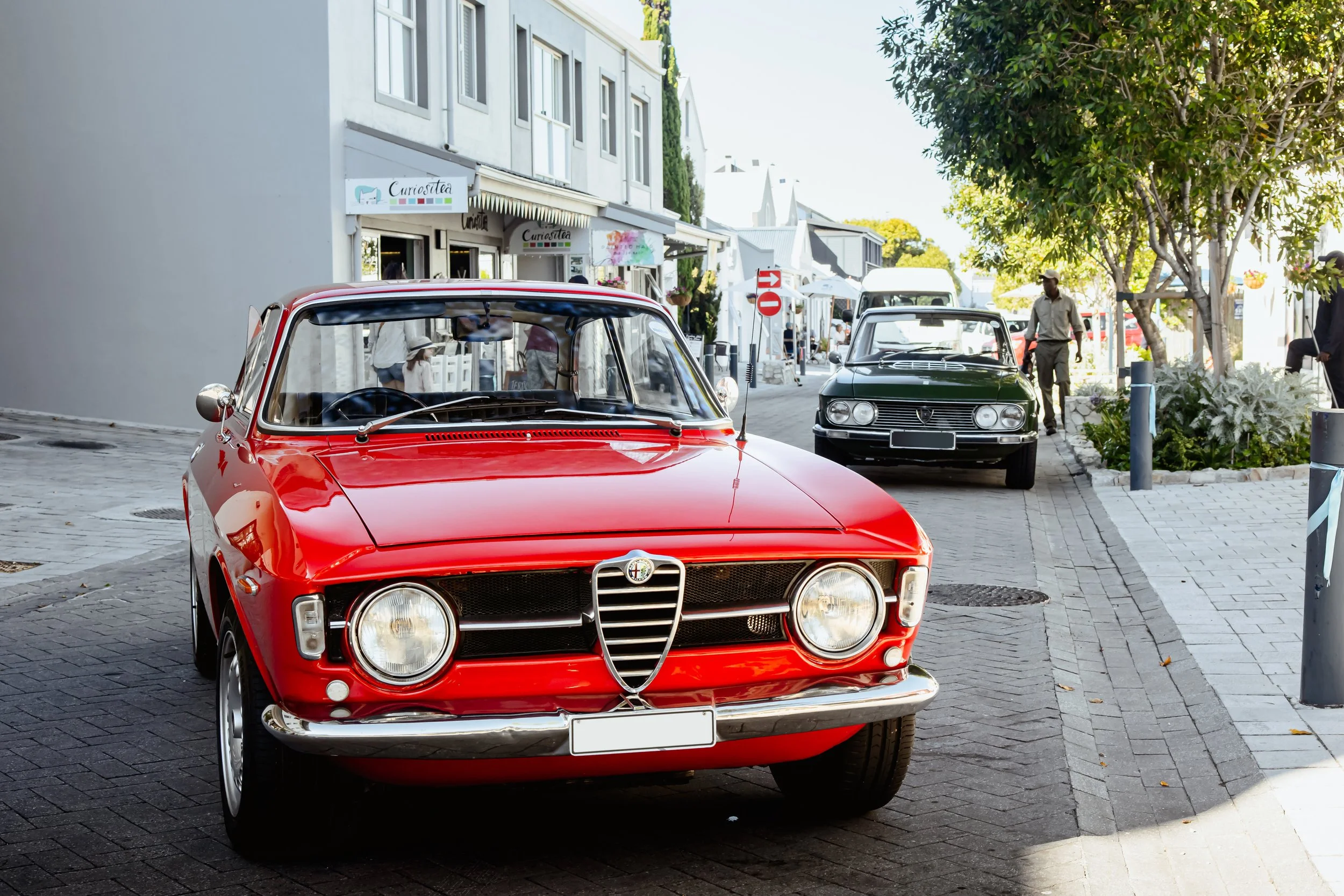 A red vintage Alfa Romeo car parked on a city street with other cars and pedestrians, alongside storefronts and trees.