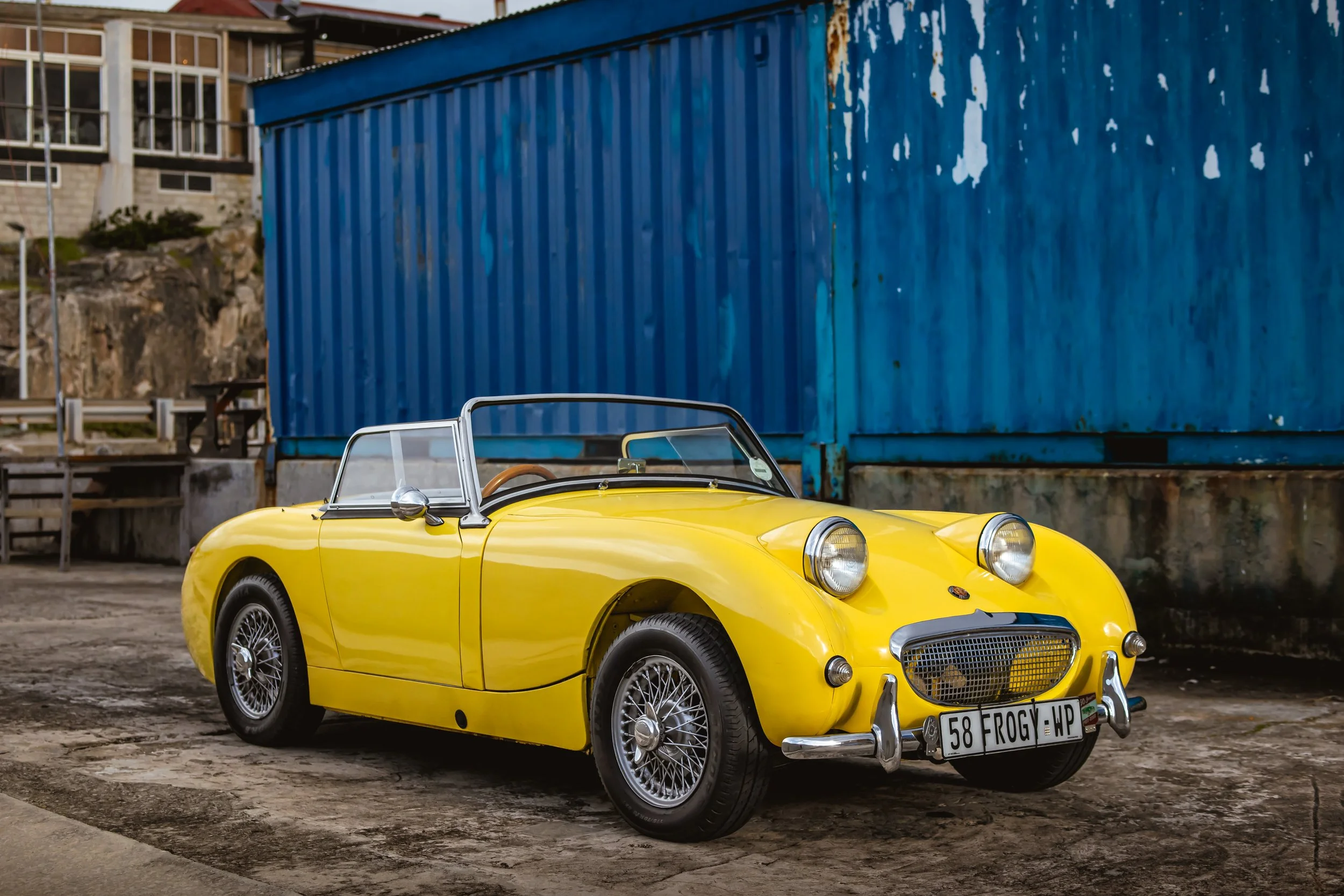 A vintage yellow Austin Healey convertible sports car parked on a dirt surface in front of a weathered blue shipping container.