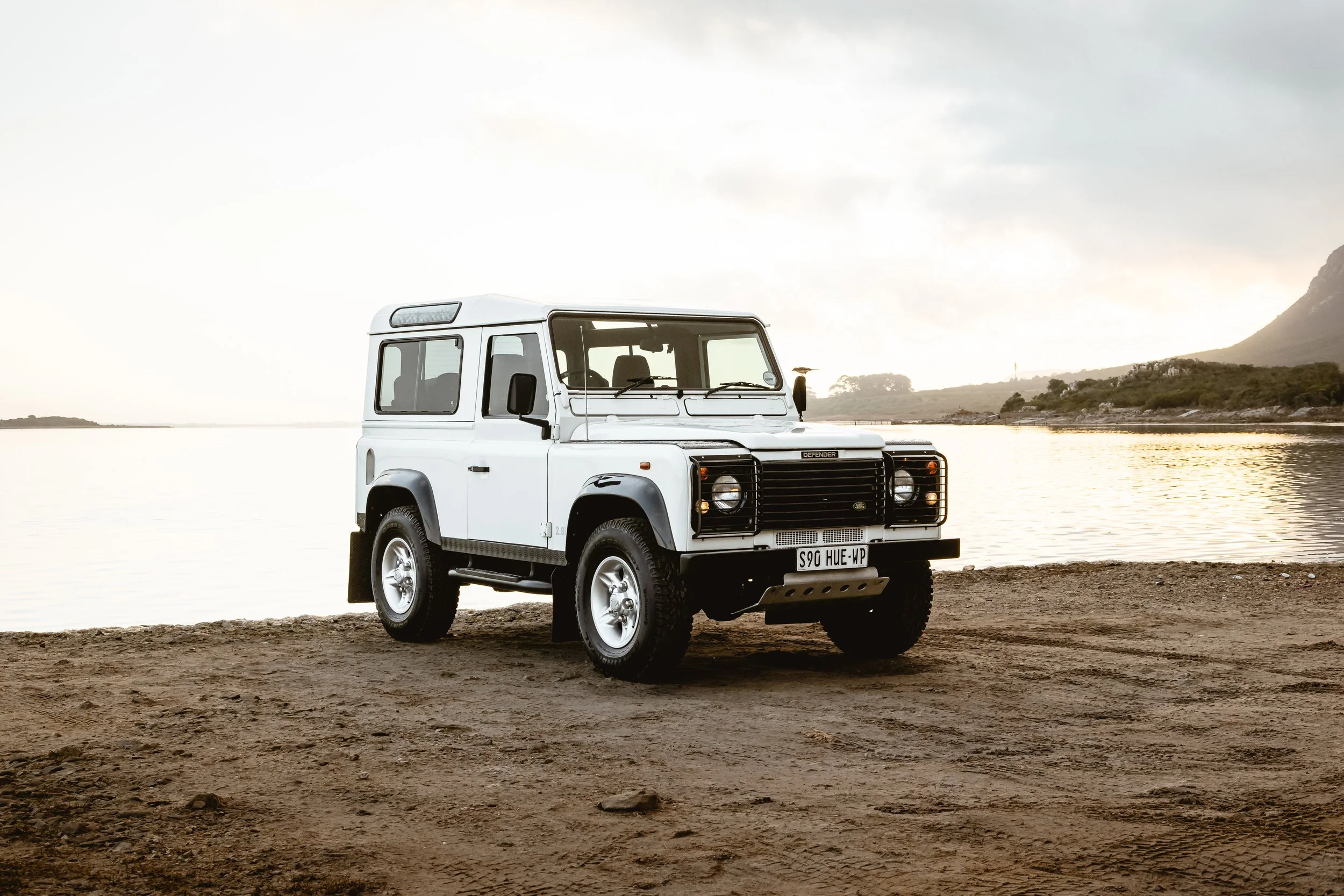 White Land Rover Defender parked on a sandy beach near a body of water with hills and a mountain in the background at sunset.