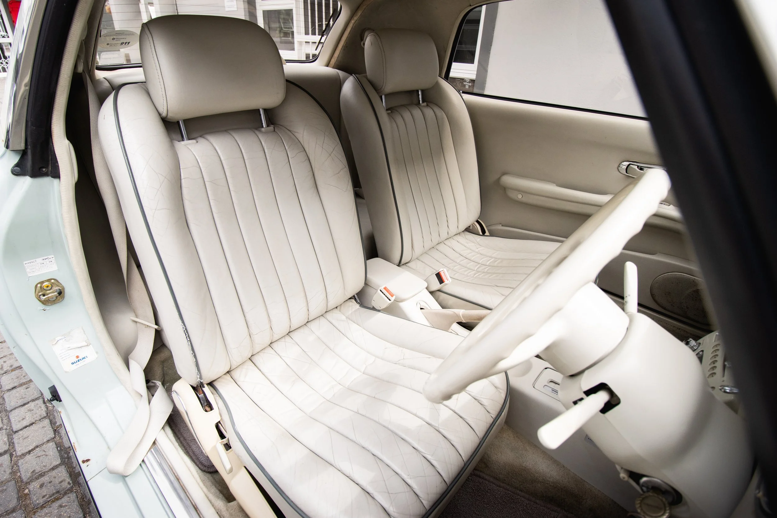 Front and back seats of a vintage beige car with fabric upholstery, a white steering wheel, and a beige door panel. The car interior looks well-maintained and clean.