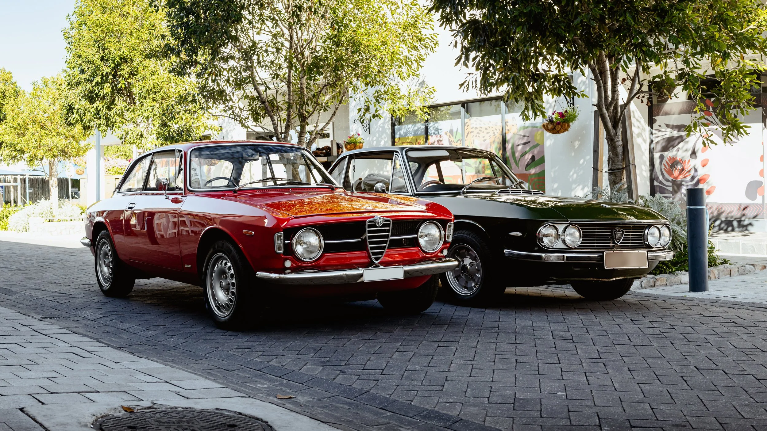 Two vintage cars, one red and one black, parked side by side on a paved street with trees and modern buildings in the background.