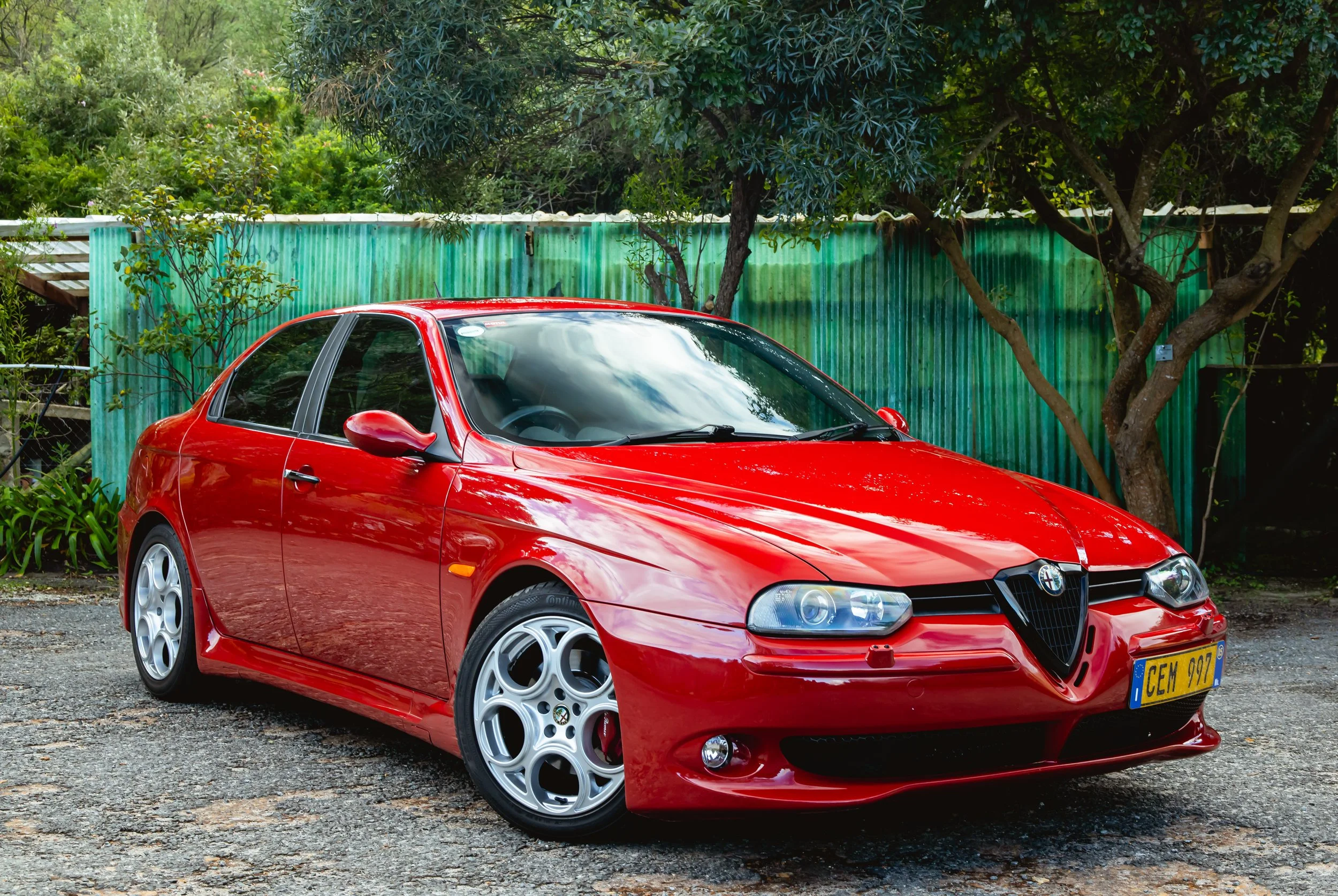 Red Alfa Romeo 159 GTA sedan parked on a gravel surface with trees and green corrugated metal fence in the background.