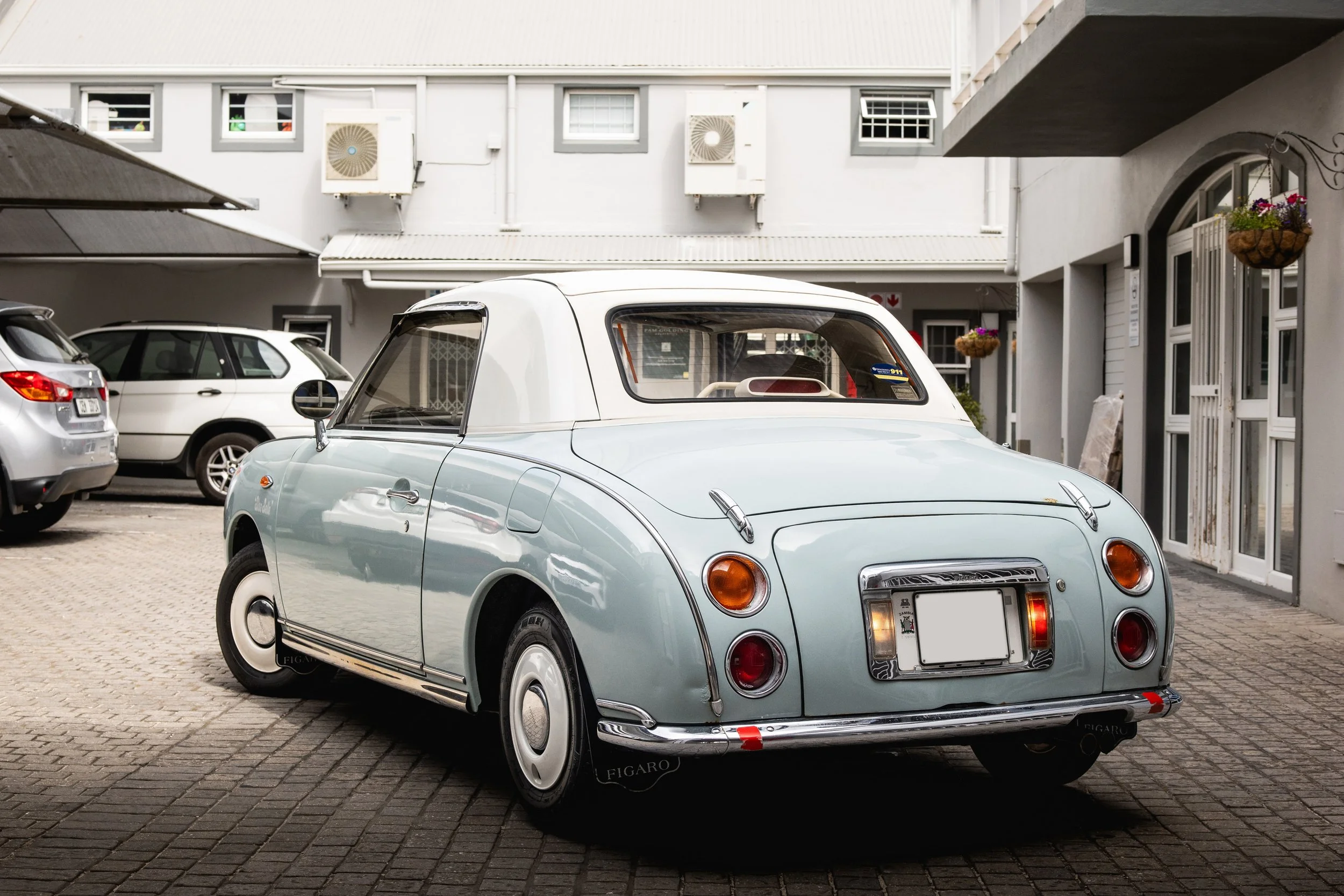 Light blue vintage convertible car parked on a brick pavement in an outdoor parking lot with other cars and a white building in the background.